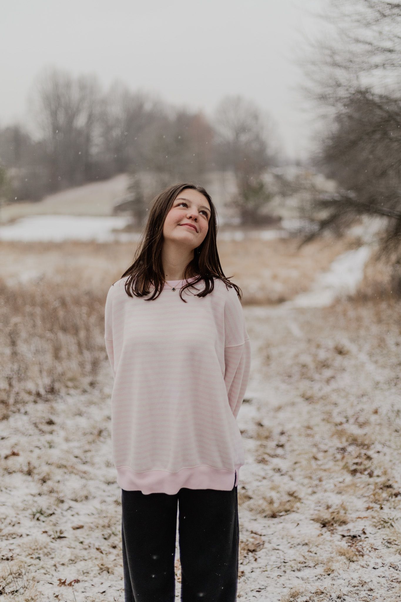 A winter portrait of someone in a pink sweater looking up at falling snow.