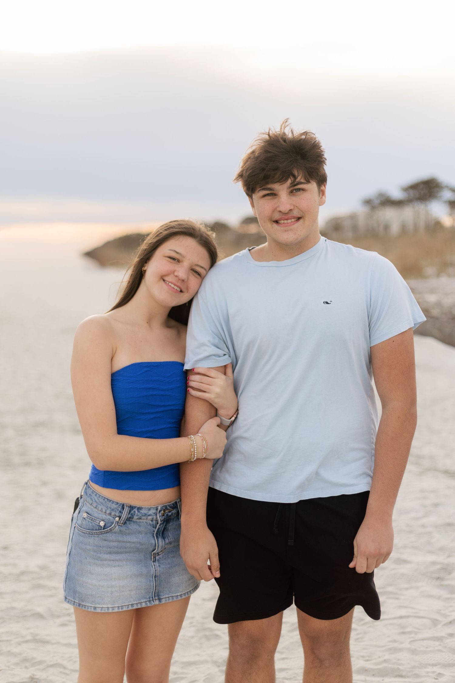 A couple poses together on a white sandy beach at sunset wearing casual summer outfits.