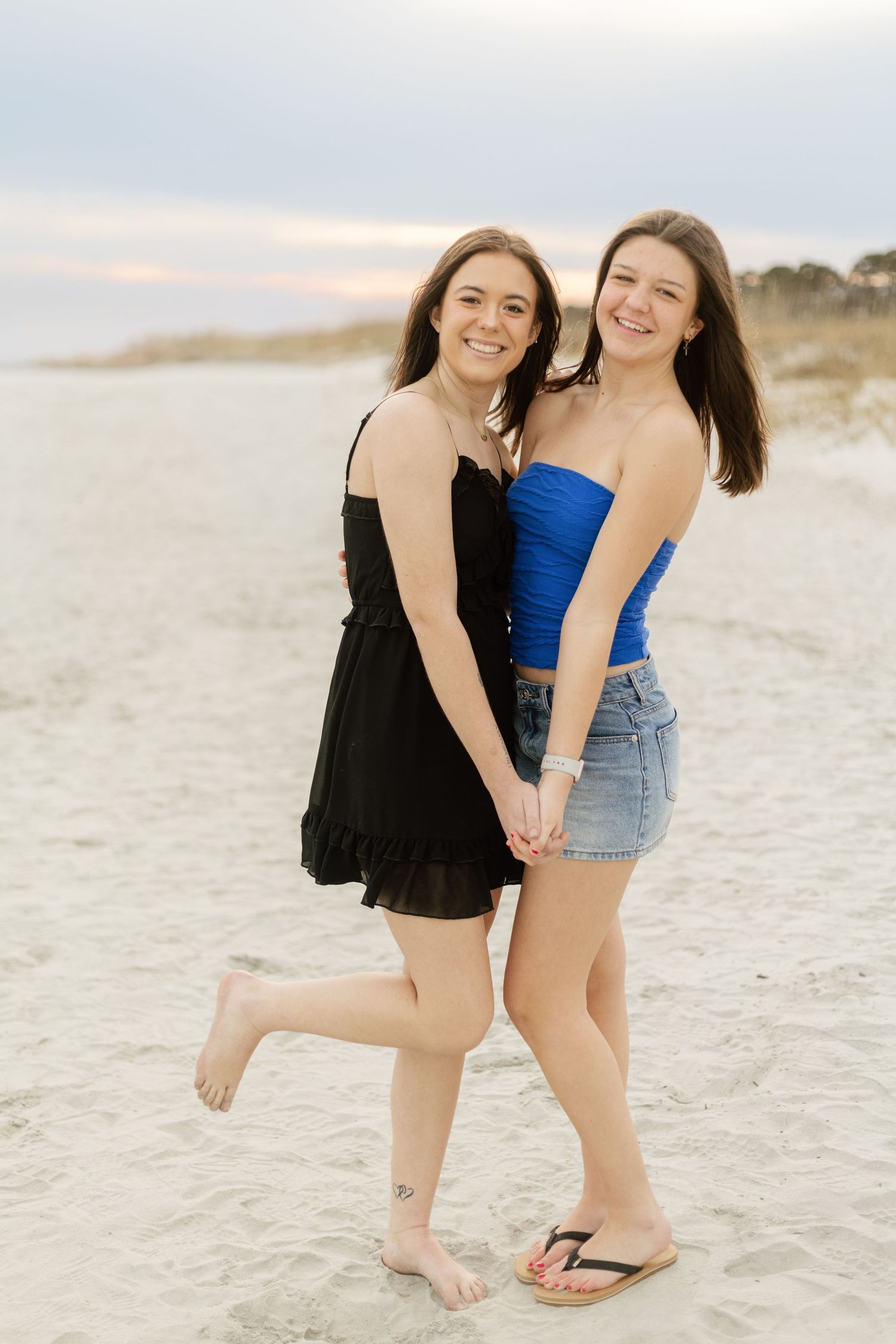 Two young women smiling and posing together on a sandy beach at sunset, one in black dress, one in blue top.