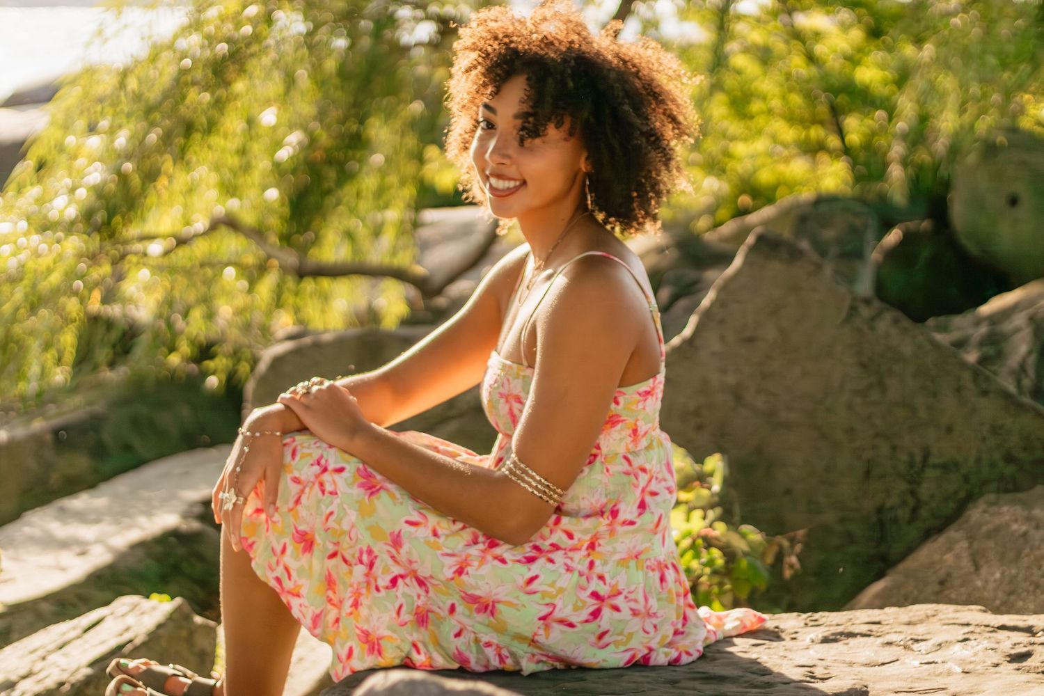 Senior in Highschool in floral dress poses on rocky terrain at Egewater beach in Cleveland OH during sunset photo session.