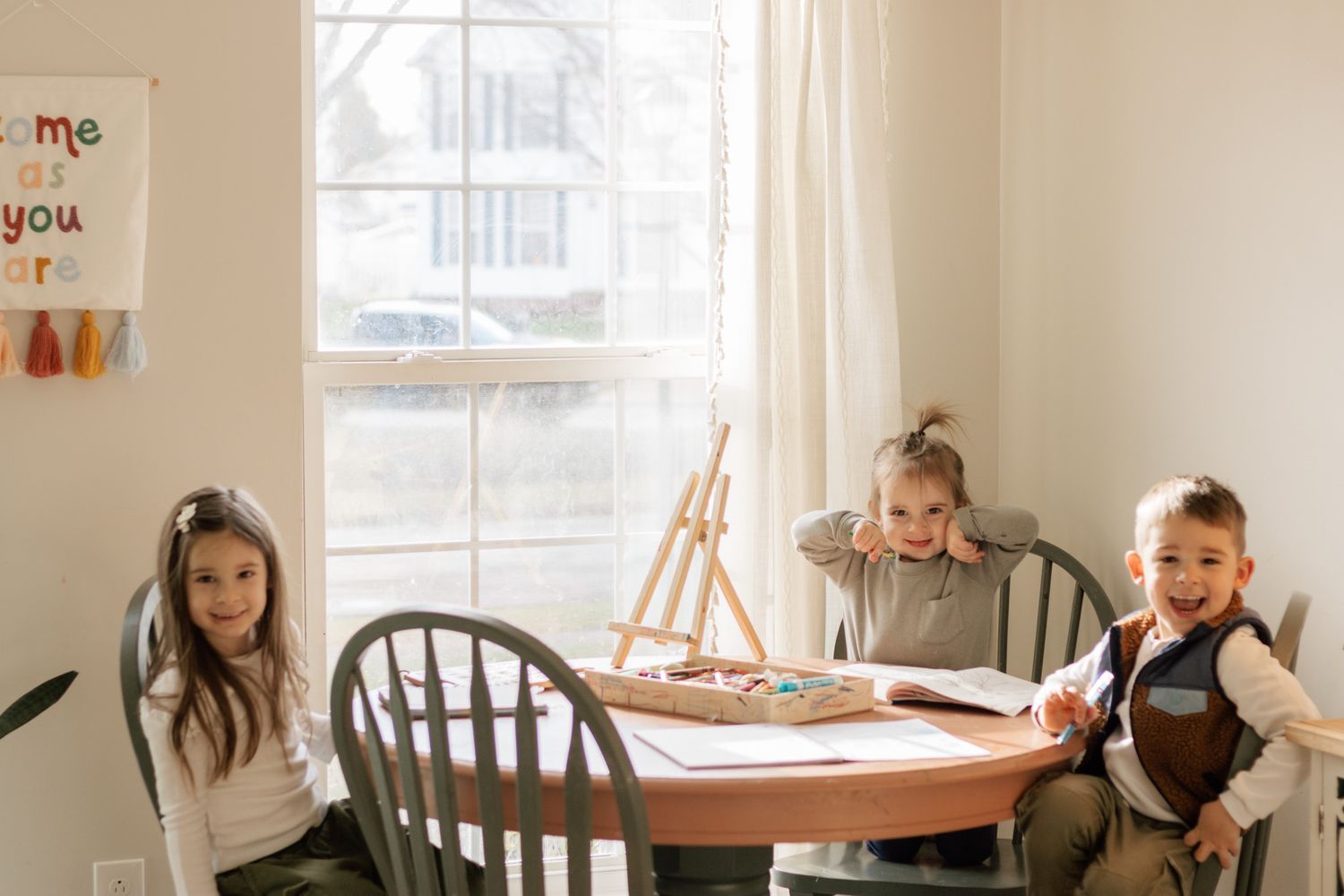 Children sitting at round wooden table near window with art supplies.