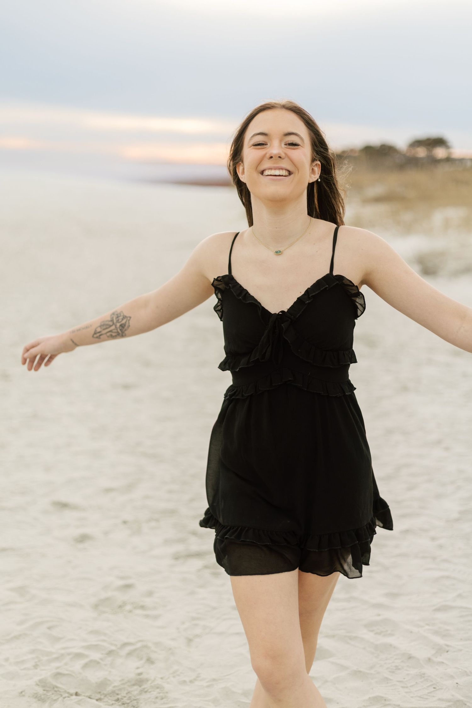 A joyful person in a black ruffle dress spins with arms outstretched on a beach at sunset.
