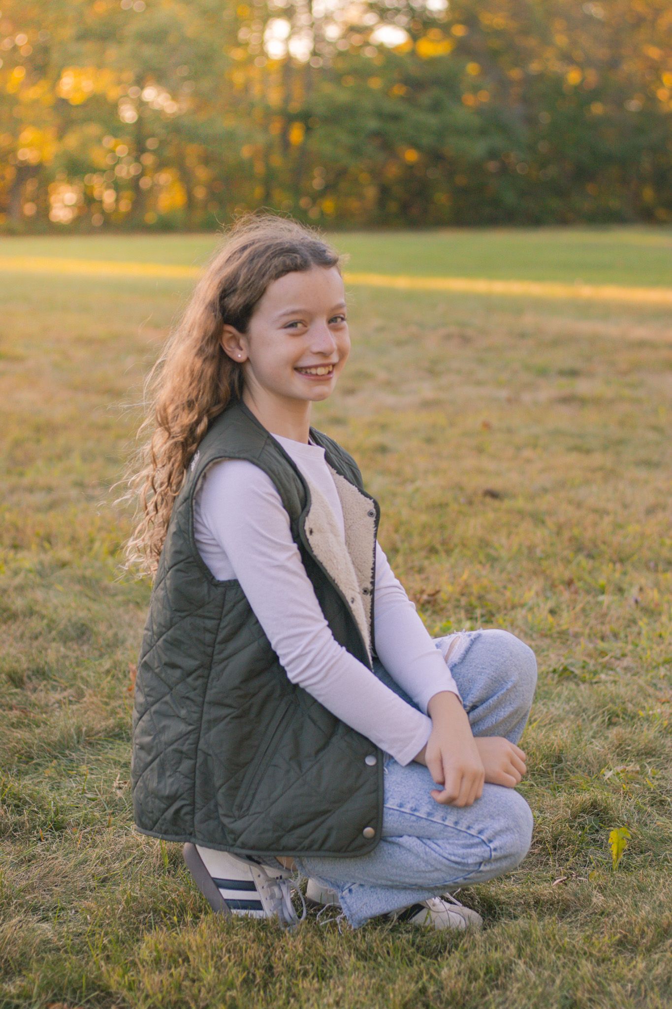 Young girl in quilted vest sits casually on grass during autumn photoshoot.