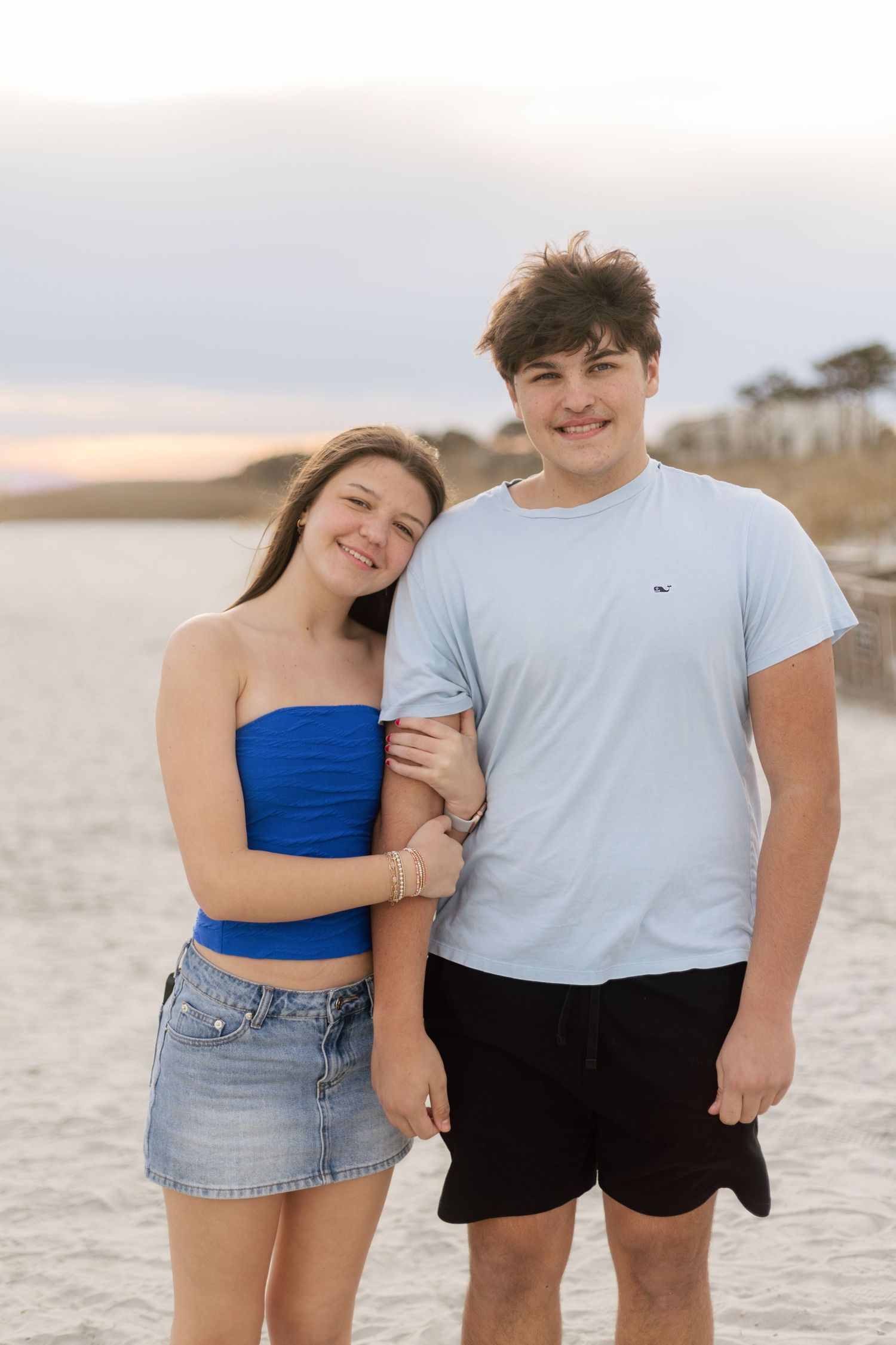 A couple in casual summer attire poses together on a sandy beach at sunset.