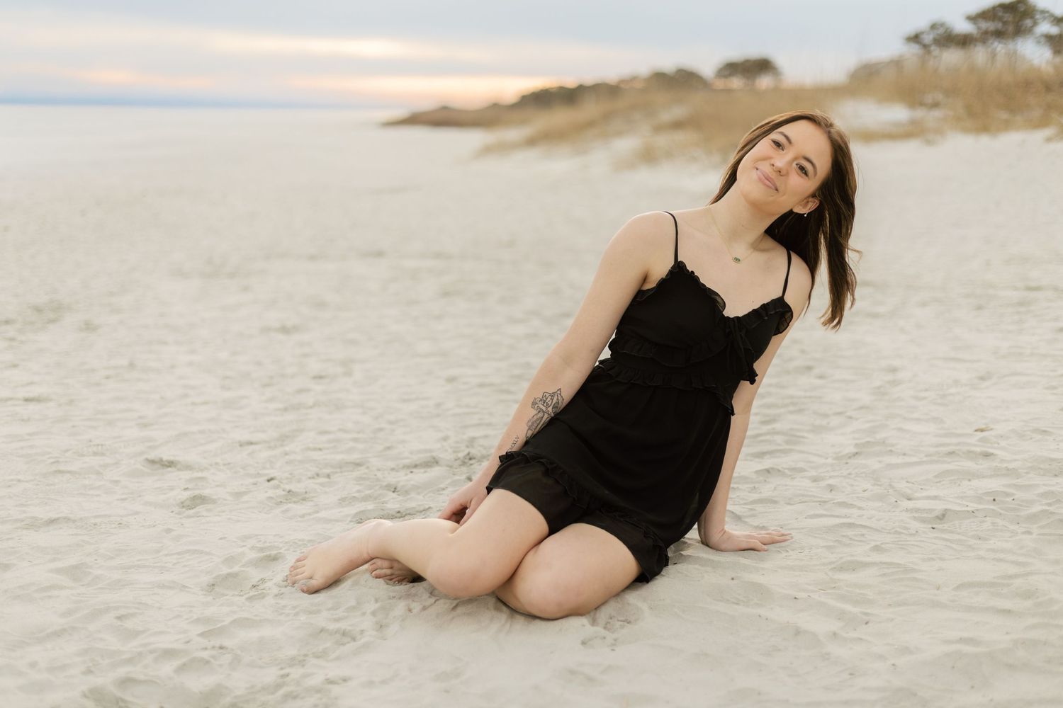 Woman in black dress poses gracefully on sandy beach during sunset photoshoot.
