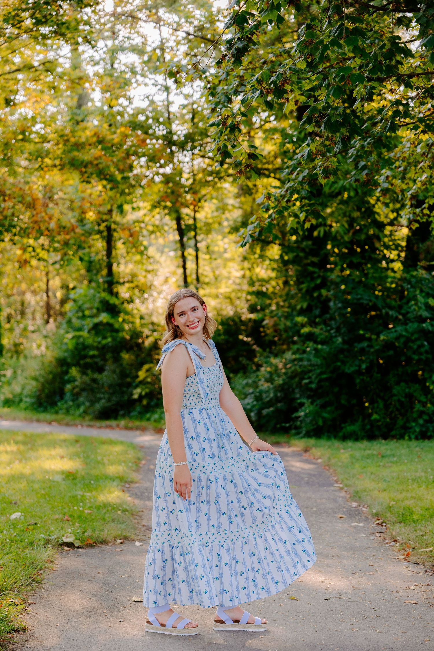 Whimsical outdoor photoshoot showing twirls and poses in a flowing blue floral dress on a tree-lined path.