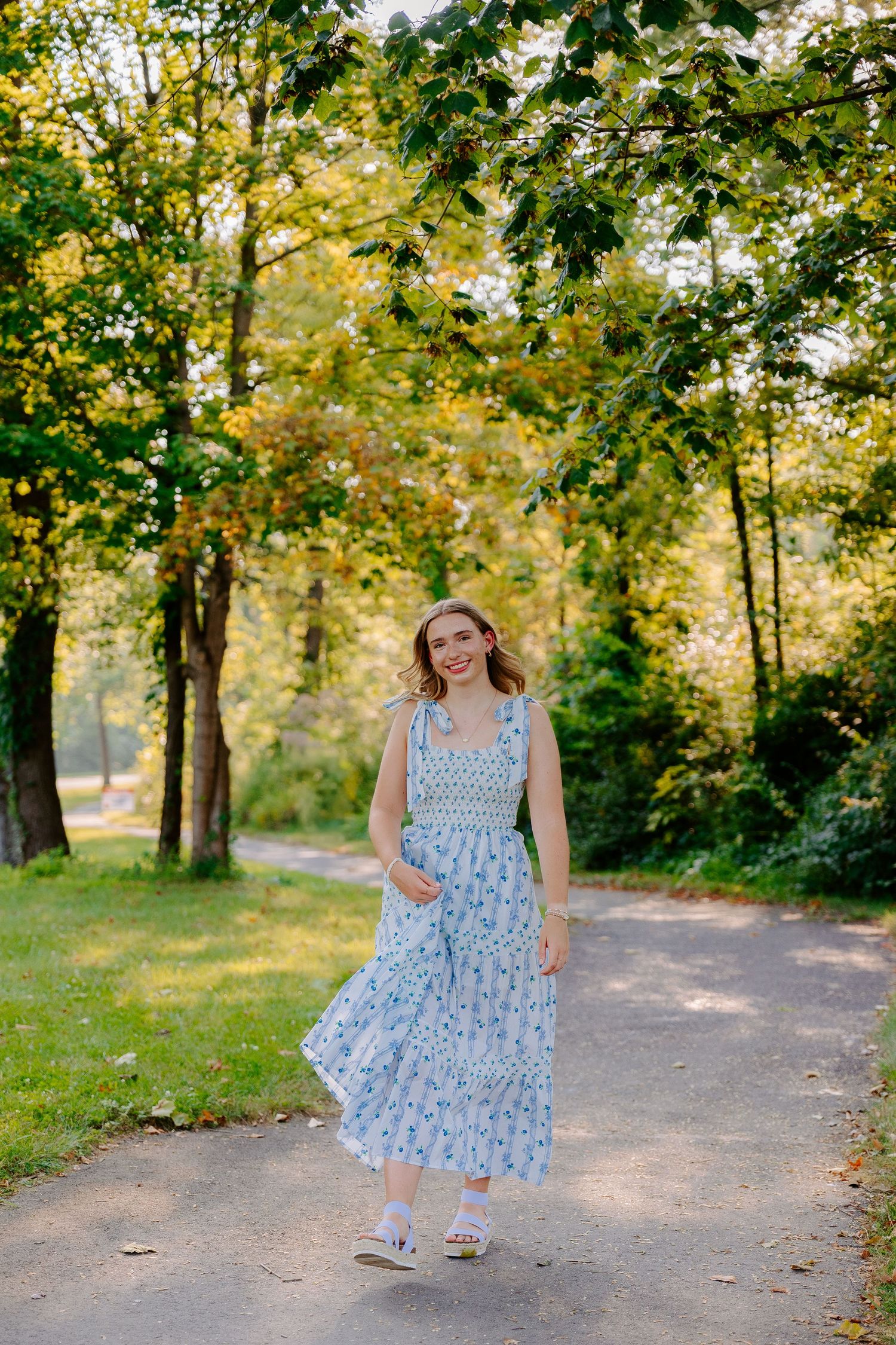 A floral blue summer dress twirls in motion against a sunlit wooded path background.