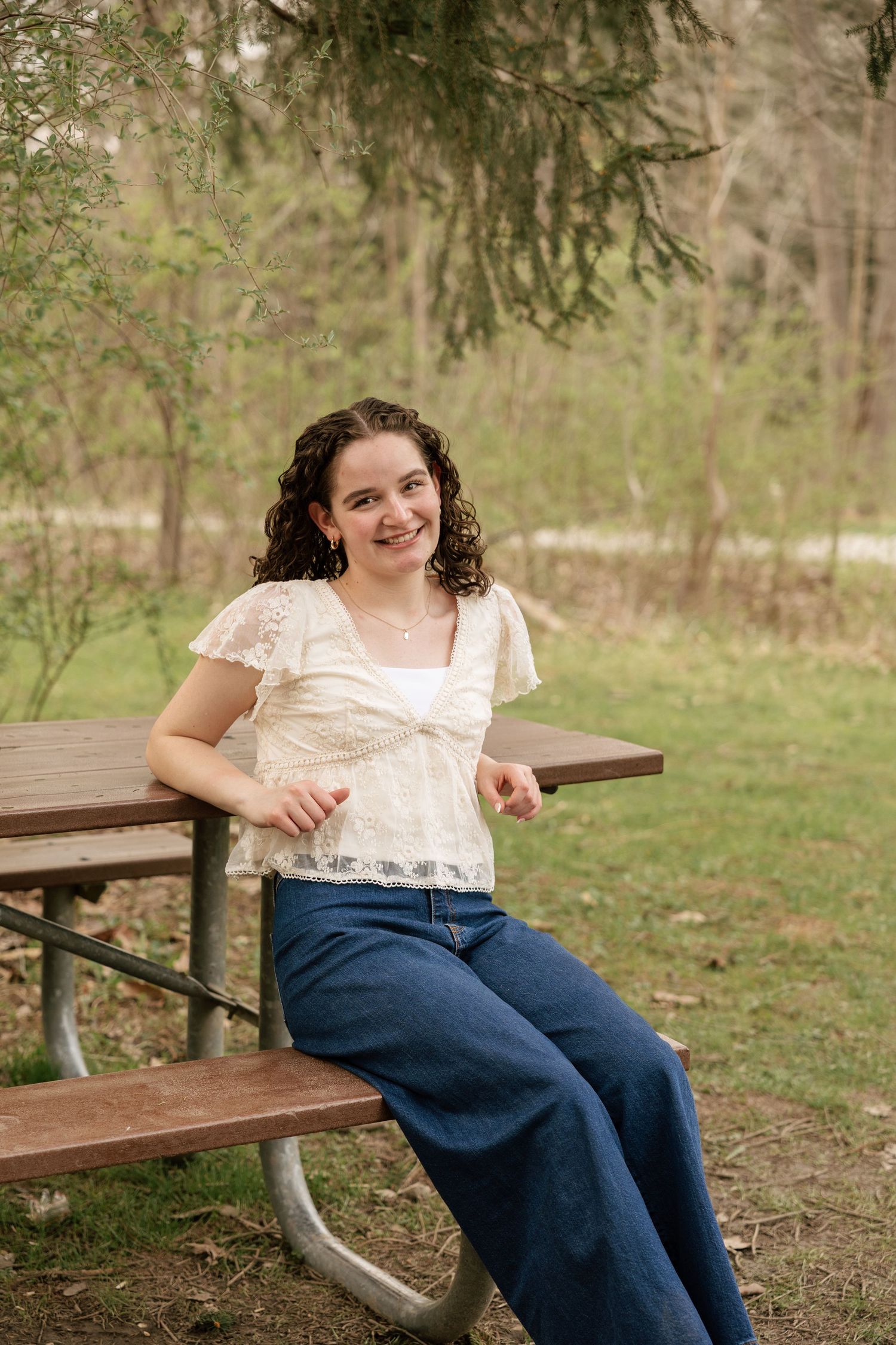 Young woman with curly hair smiles while sitting at a picnic table outdoors, wearing a lace top and wide-leg jeans.