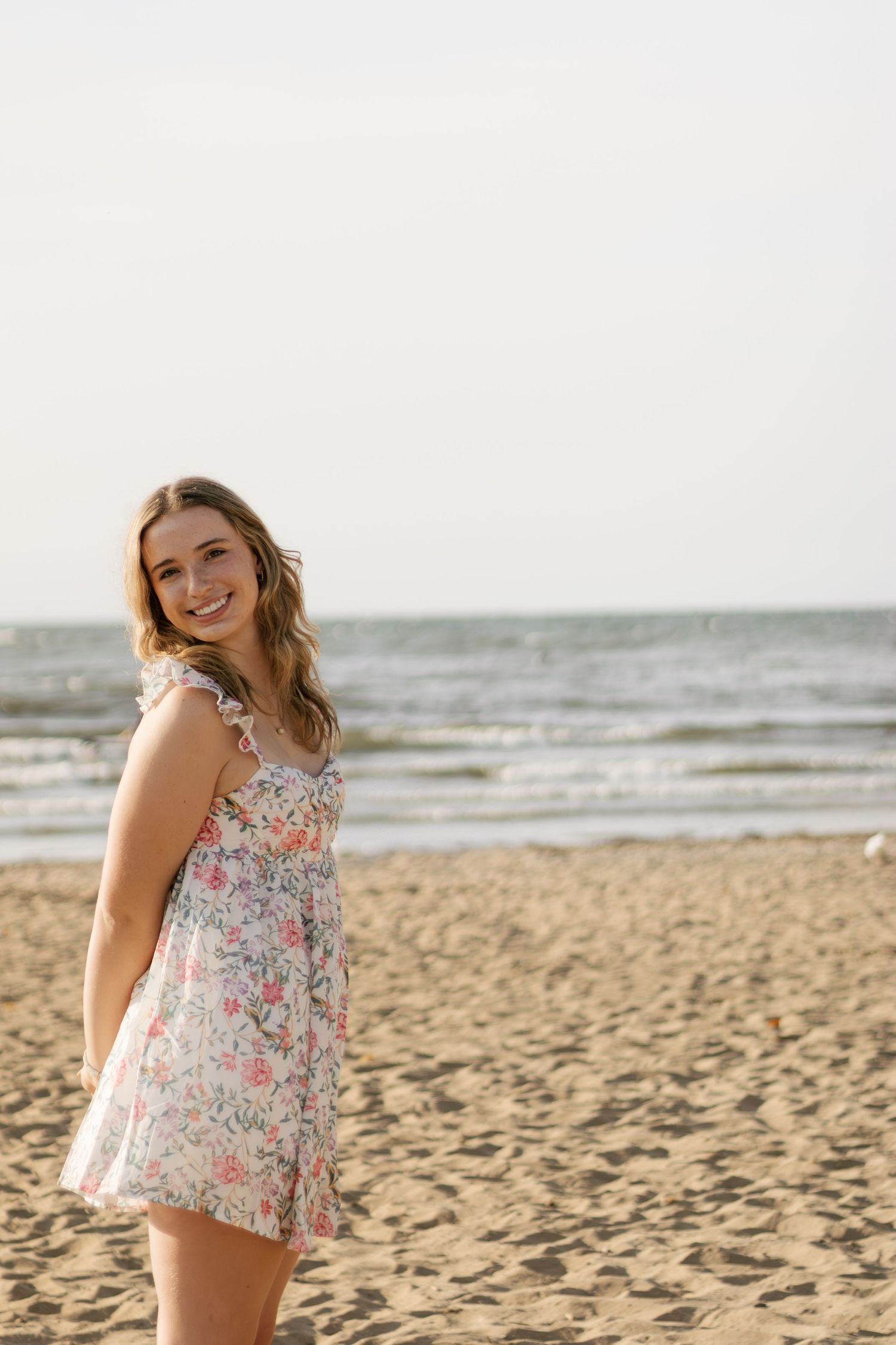 Senior in floral top poses playfully on sandy beach at sunset at edgewater beach in Cleveland, OH during her senior photos