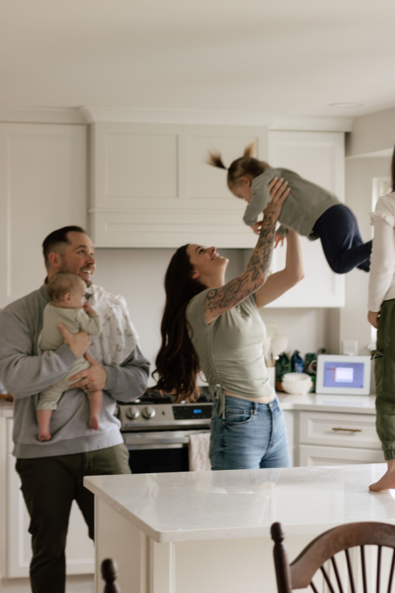 Family playing and lifting children in modern white kitchen with holiday decor.