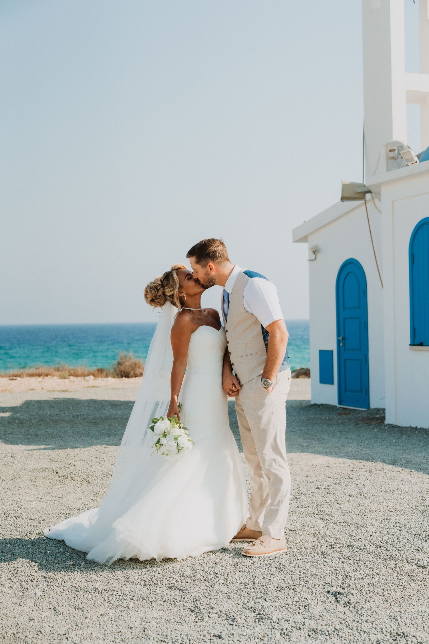 Bride and groom share a kiss near a white Greek chapel with blue doors by the sea on their wedding day.
