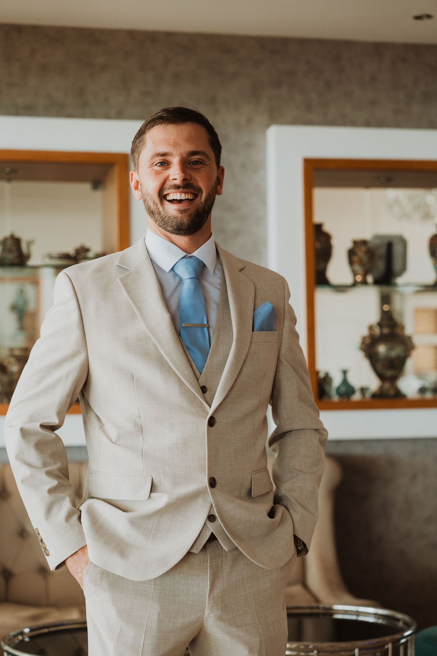 Smiling groom in beige three-piece suit with blue tie and pocket square, standing confidently indoors.