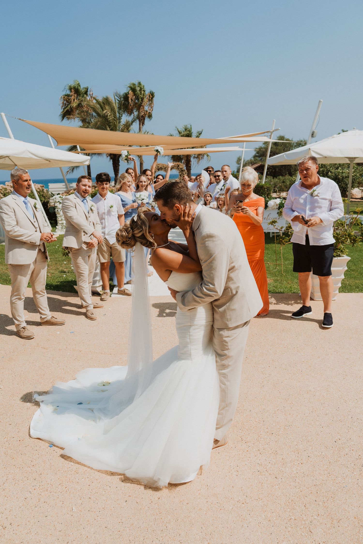 Bride and groom share a romantic dip kiss at outdoor beach wedding ceremony with guests watching.