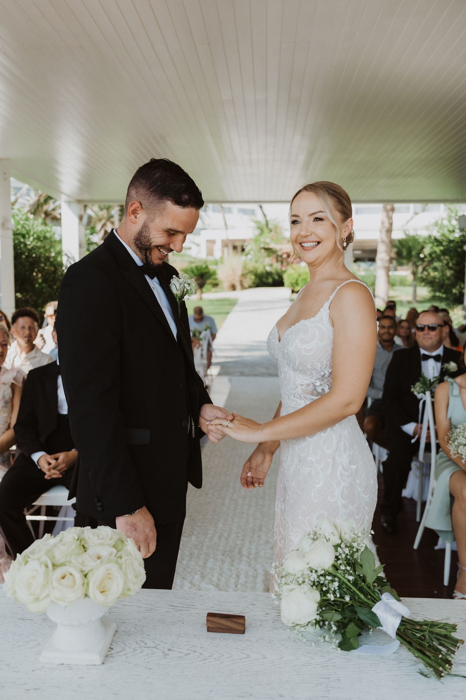 Couple exchanges rings at wedding ceremony table with white floral arrangements.