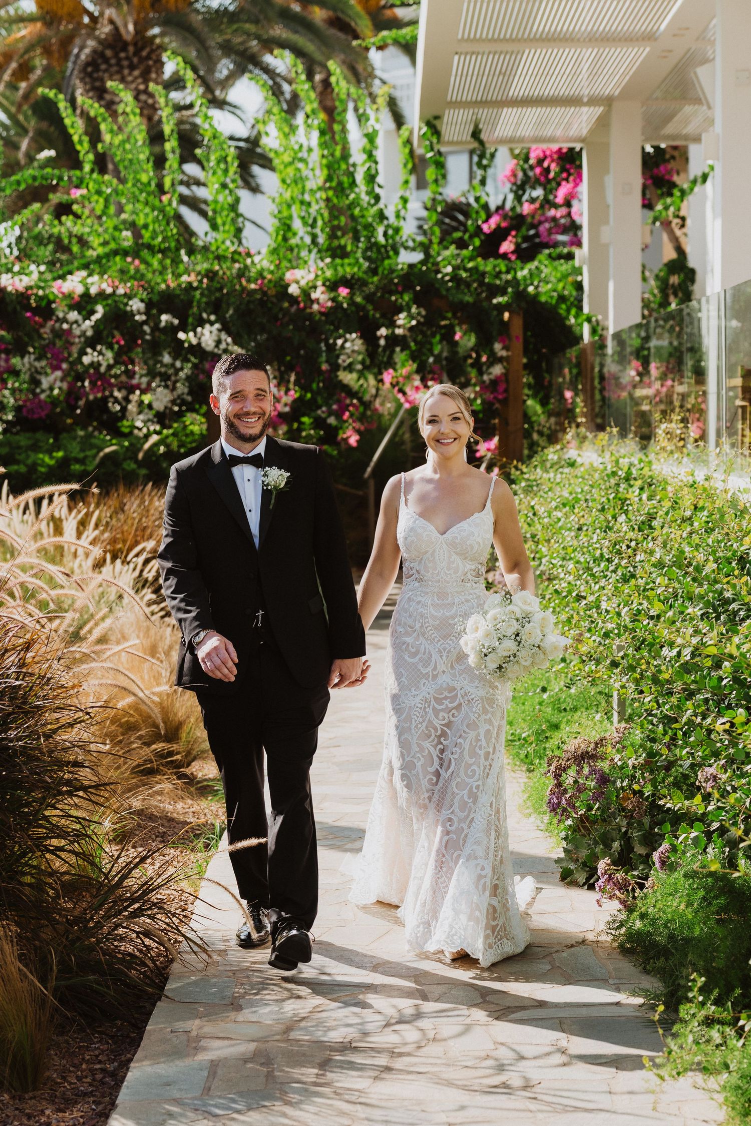 Wedding couple walks along a garden path with lush greenery and palm trees under a white pergola.