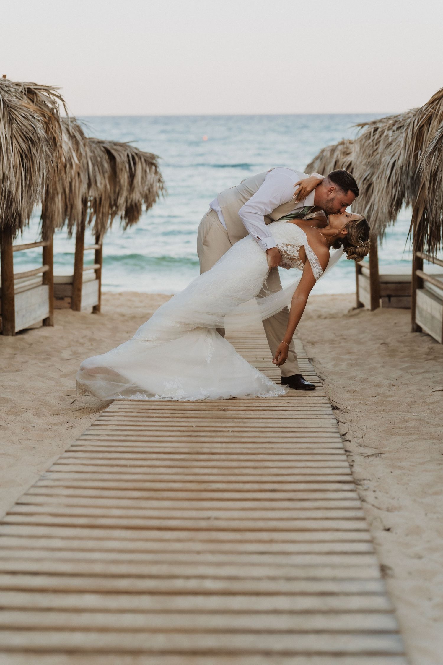 A romantic beach wedding couple shares a dramatic dip kiss on a wooden boardwalk between thatched umbrellas at sunset.