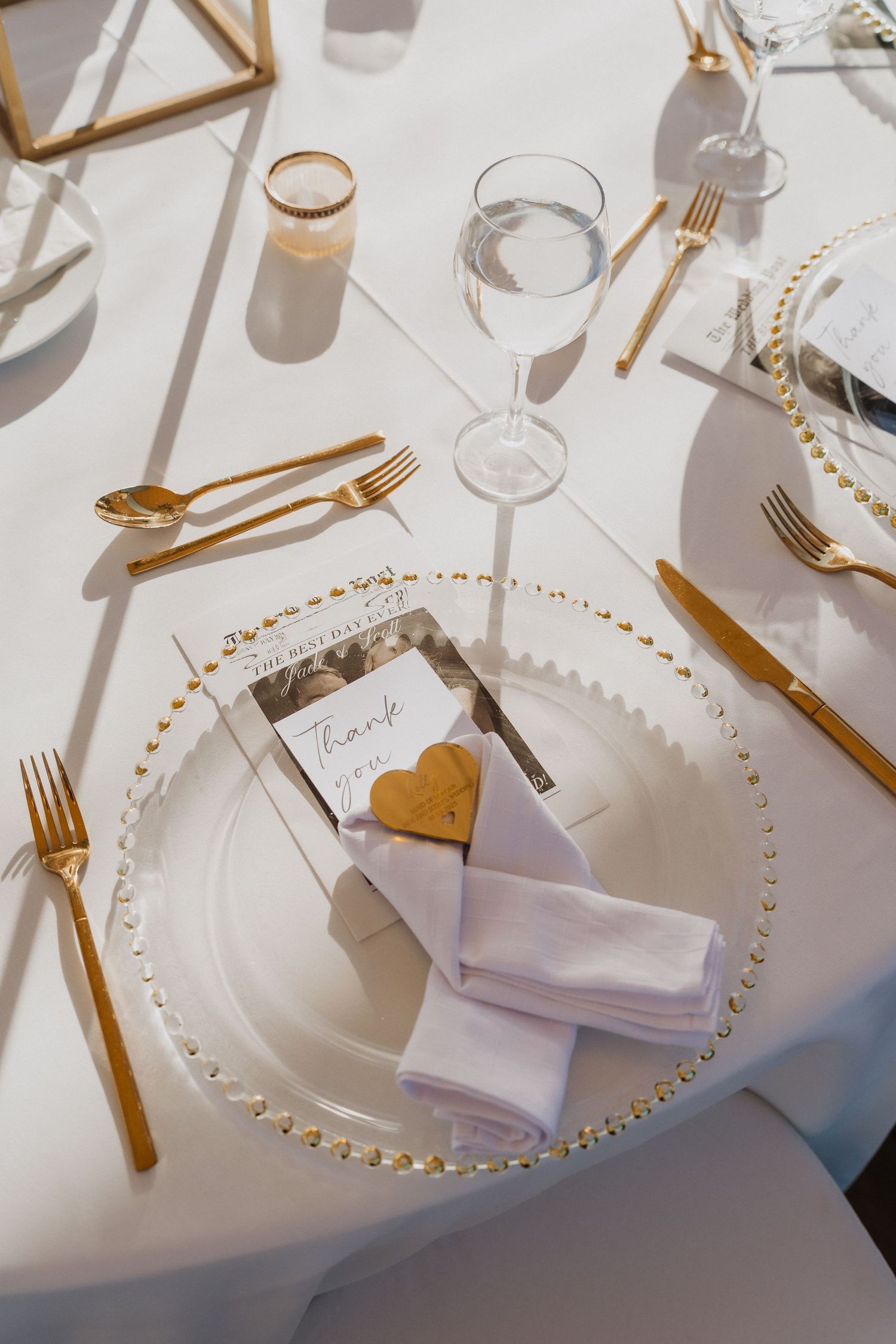 Elegant wedding place setting with gold cutlery, beaded charger plate, white napkin and heart-shaped cookie on white tablecloth.