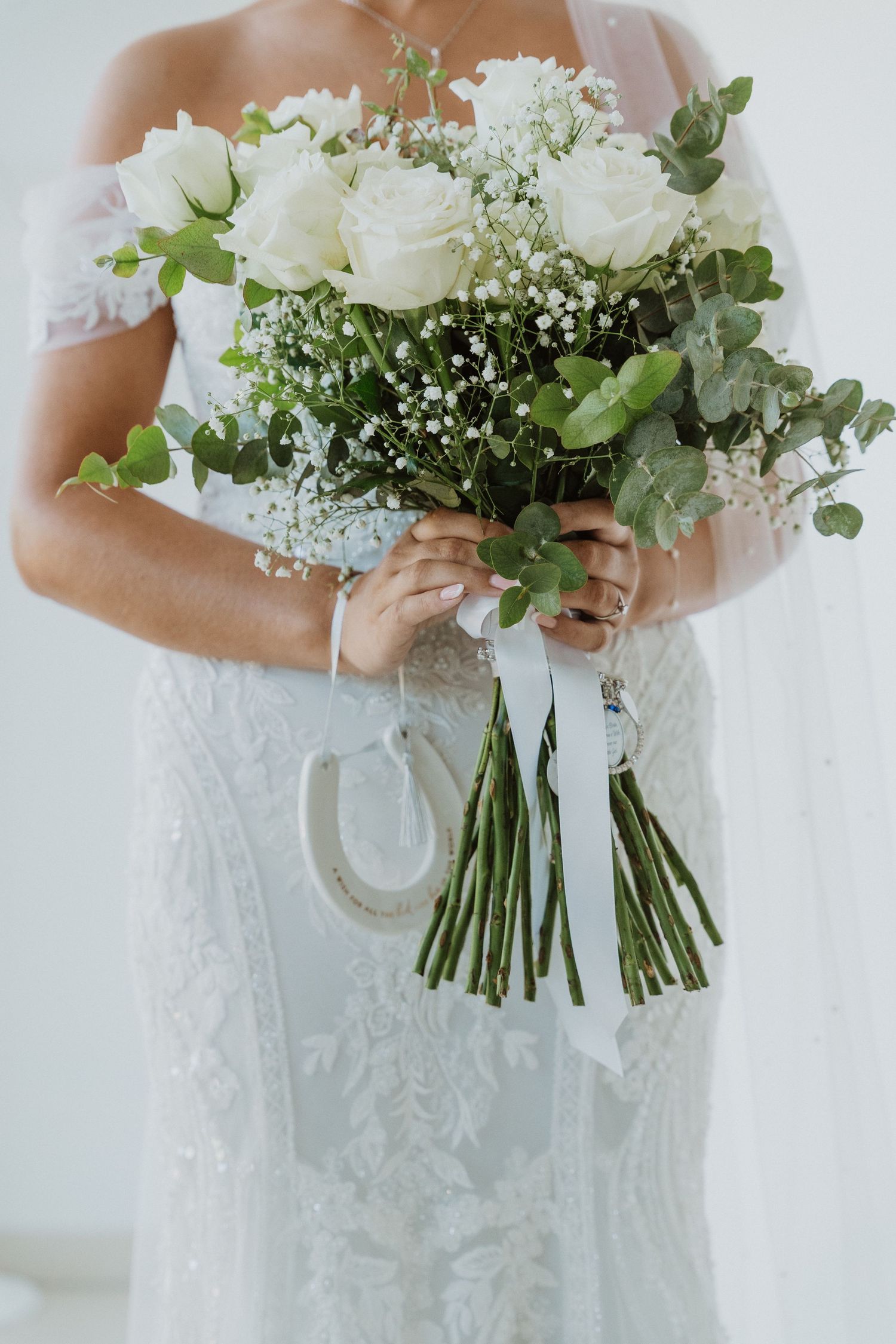 A delicate white bridal bouquet featuring roses, baby's breath and eucalyptus held against a lace wedding dress.
