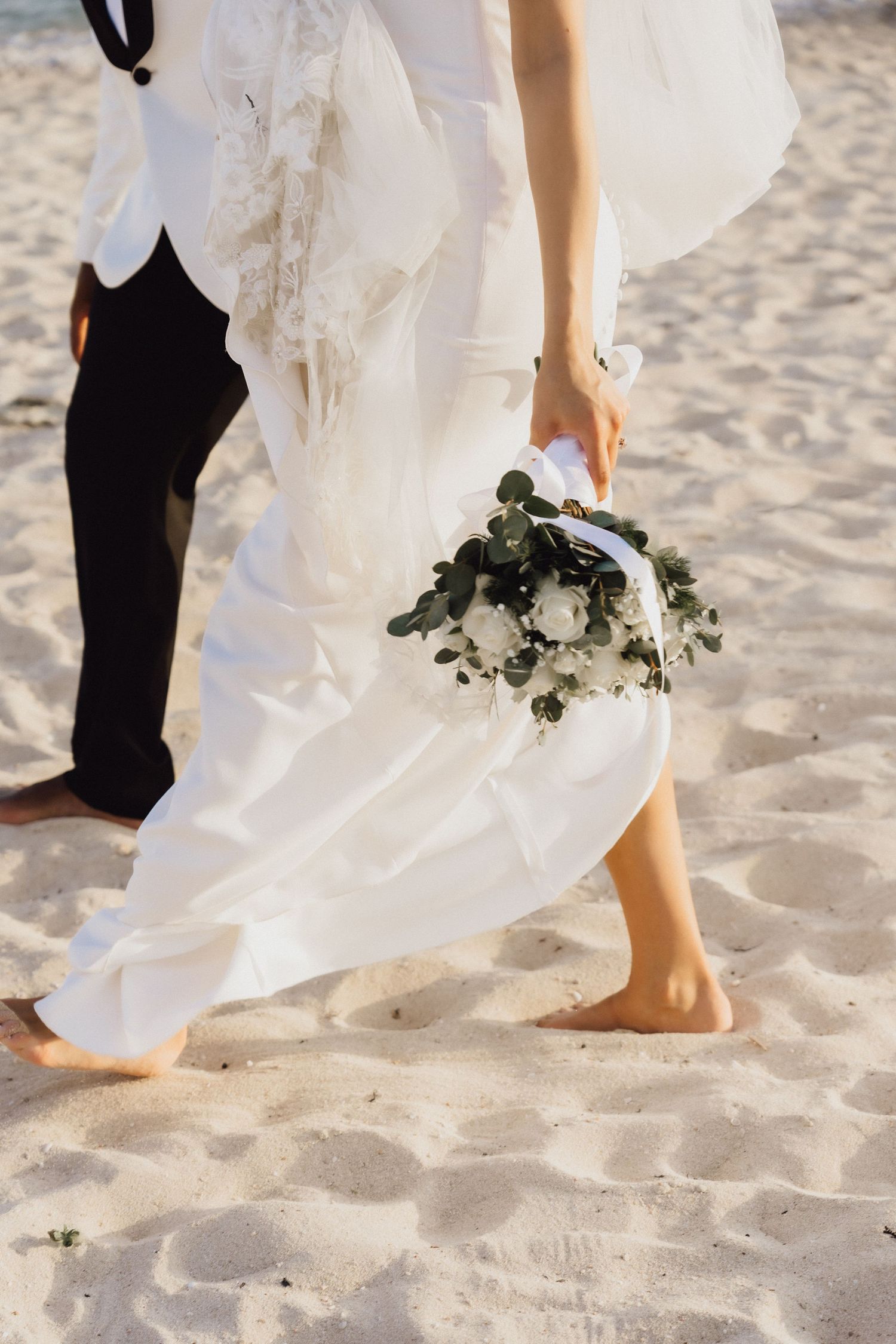 Couple's legs walking barefoot on beach sand with white wedding dress and black pants.