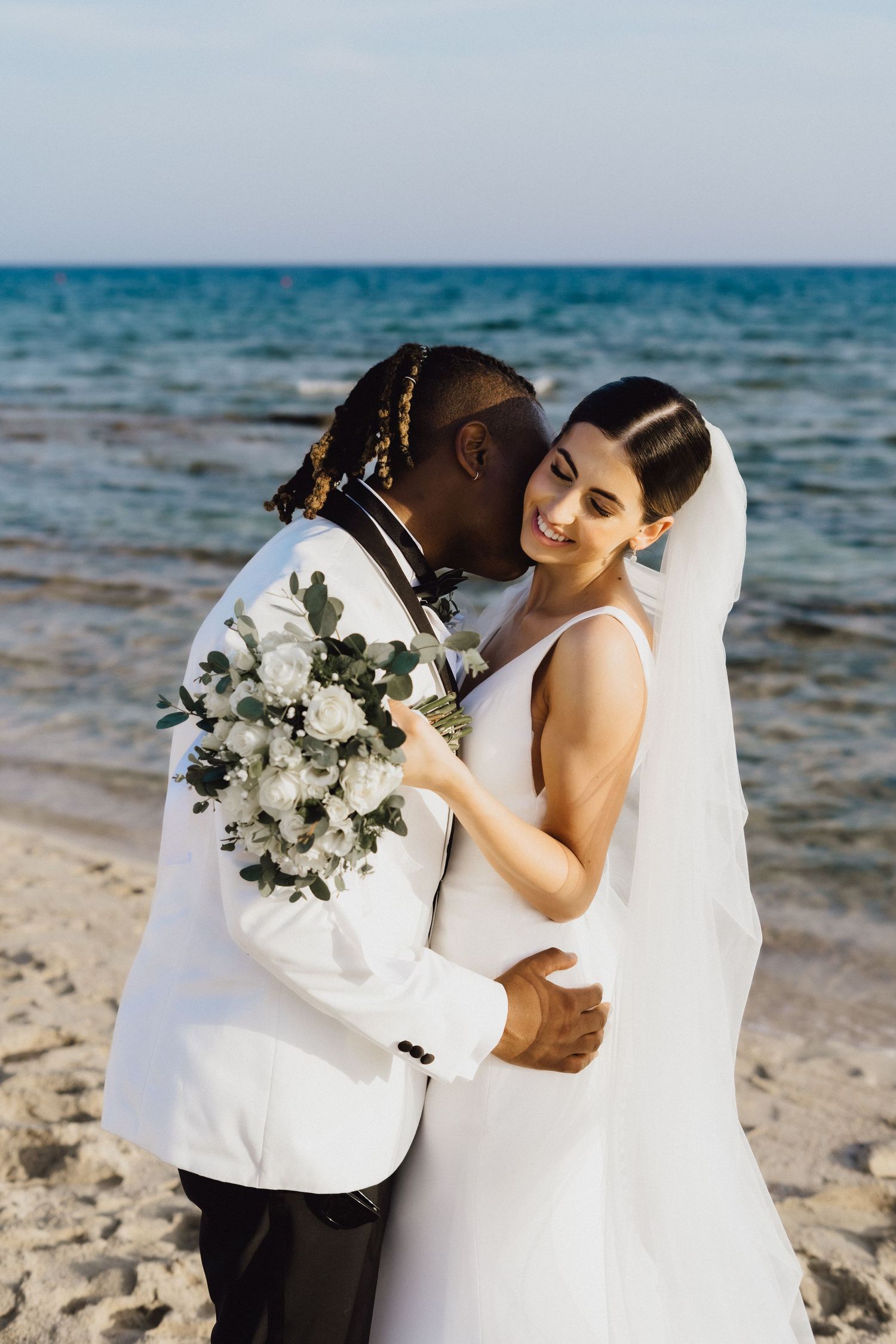 Romantic wedding photo of couple with white floral bouquet on sandy beach.