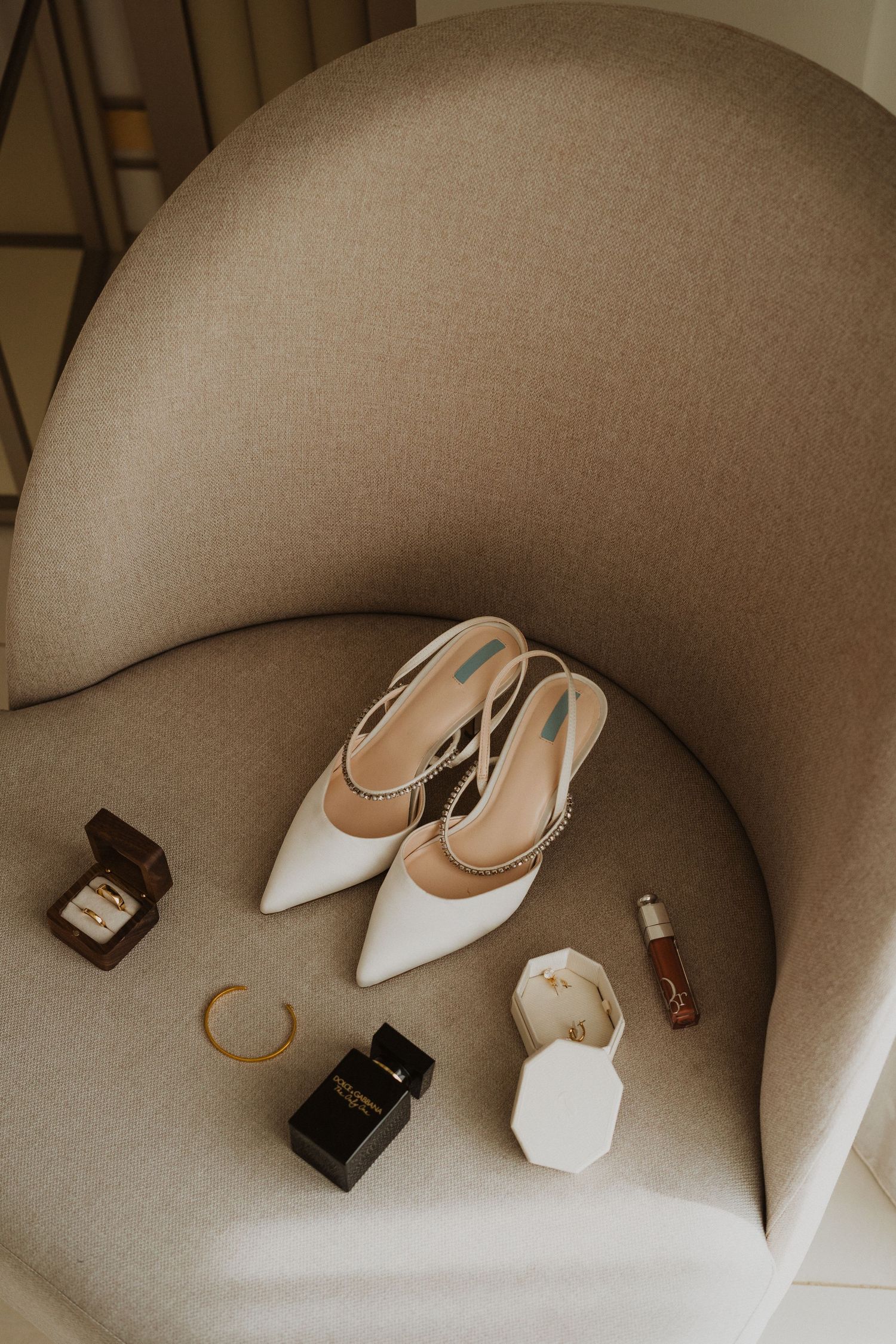 Wedding accessories including white heels, perfume, and jewelry arranged on a beige chair.