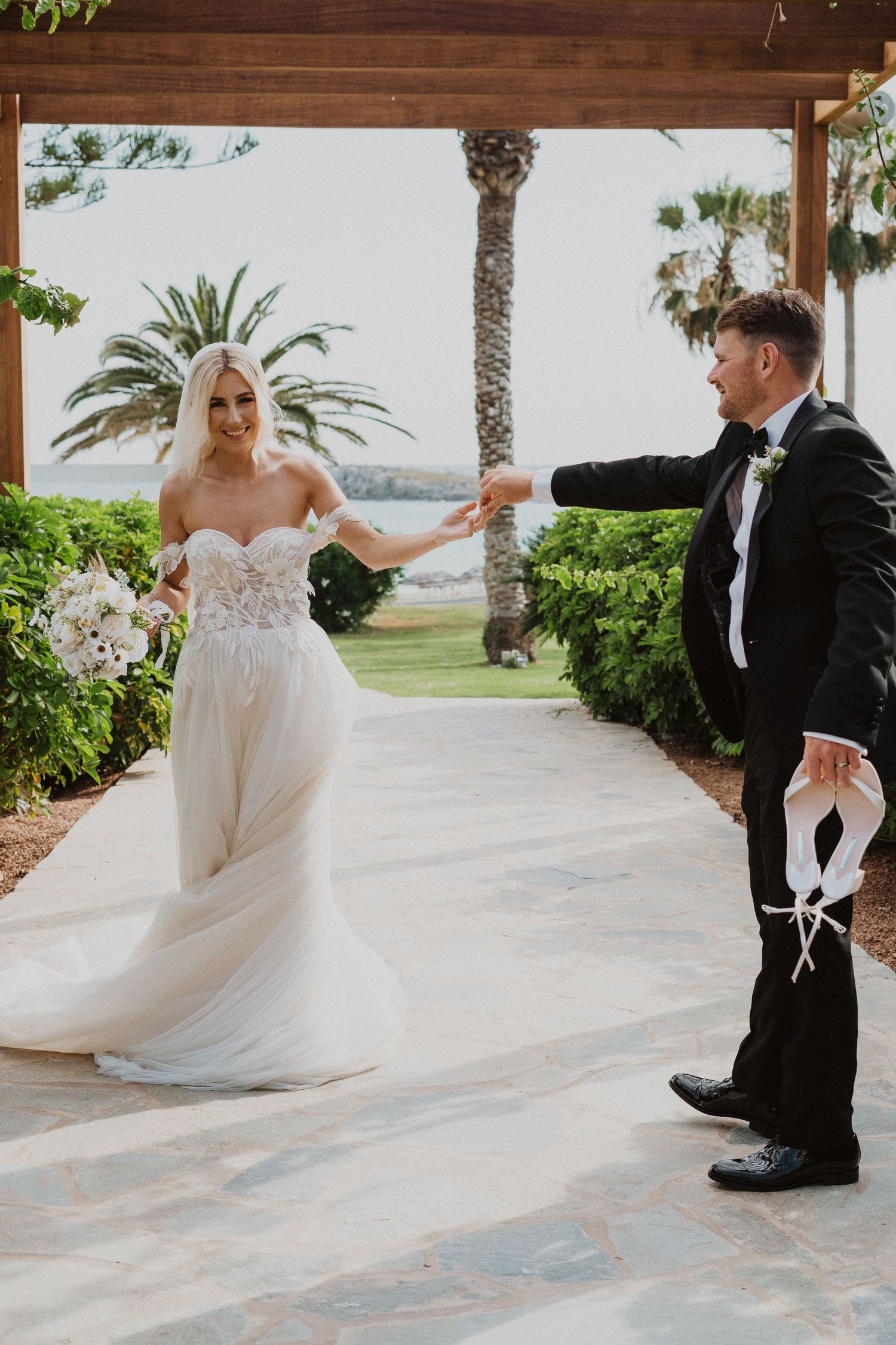 A bride in a flowing white gown and groom in black tuxedo pose playfully on a covered walkway with palm trees.
