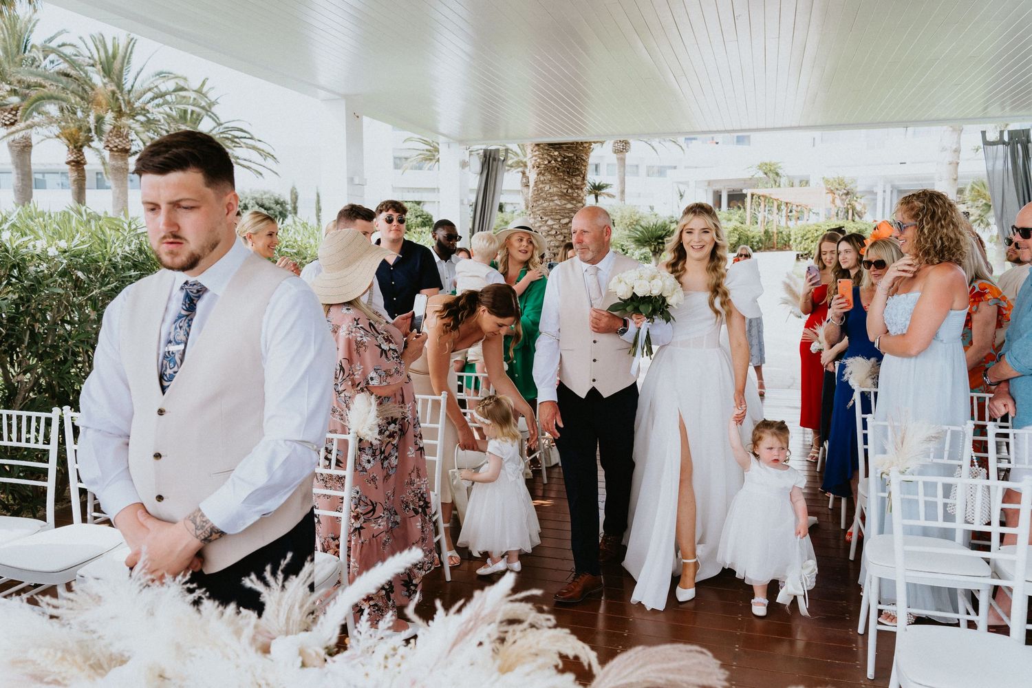 Outdoor wedding ceremony procession with white floral decor and palm trees in background.