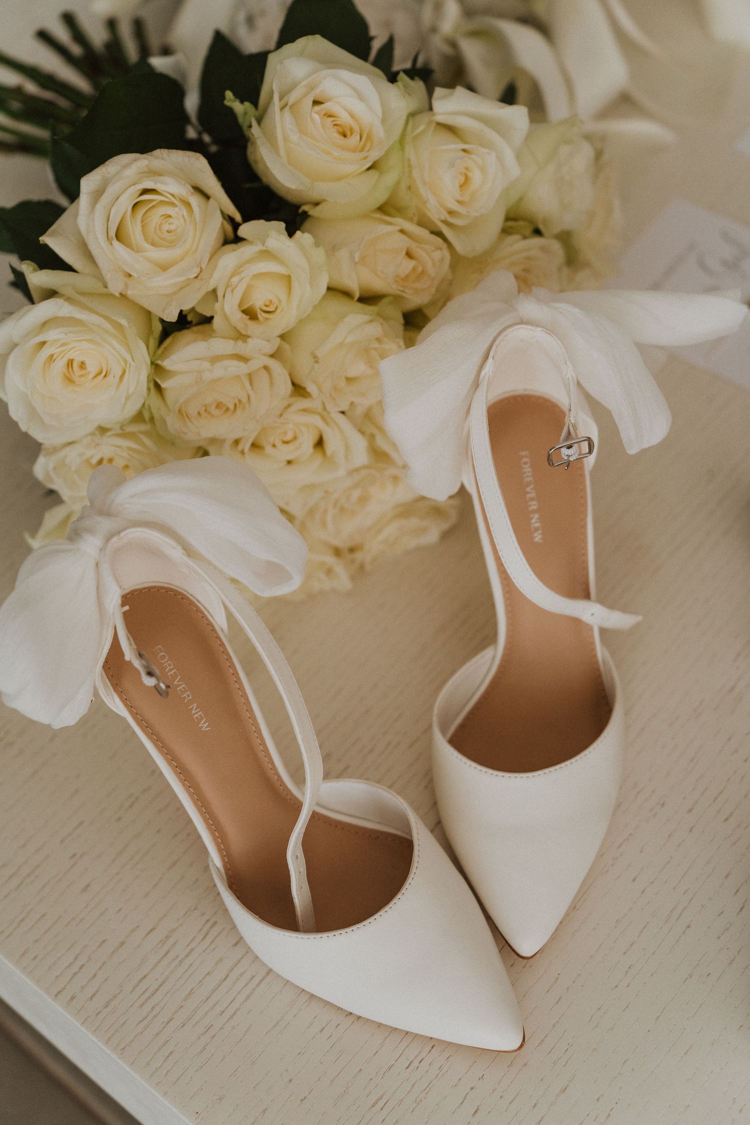 Elegant white roses and bridal heels arranged on light colored surface.