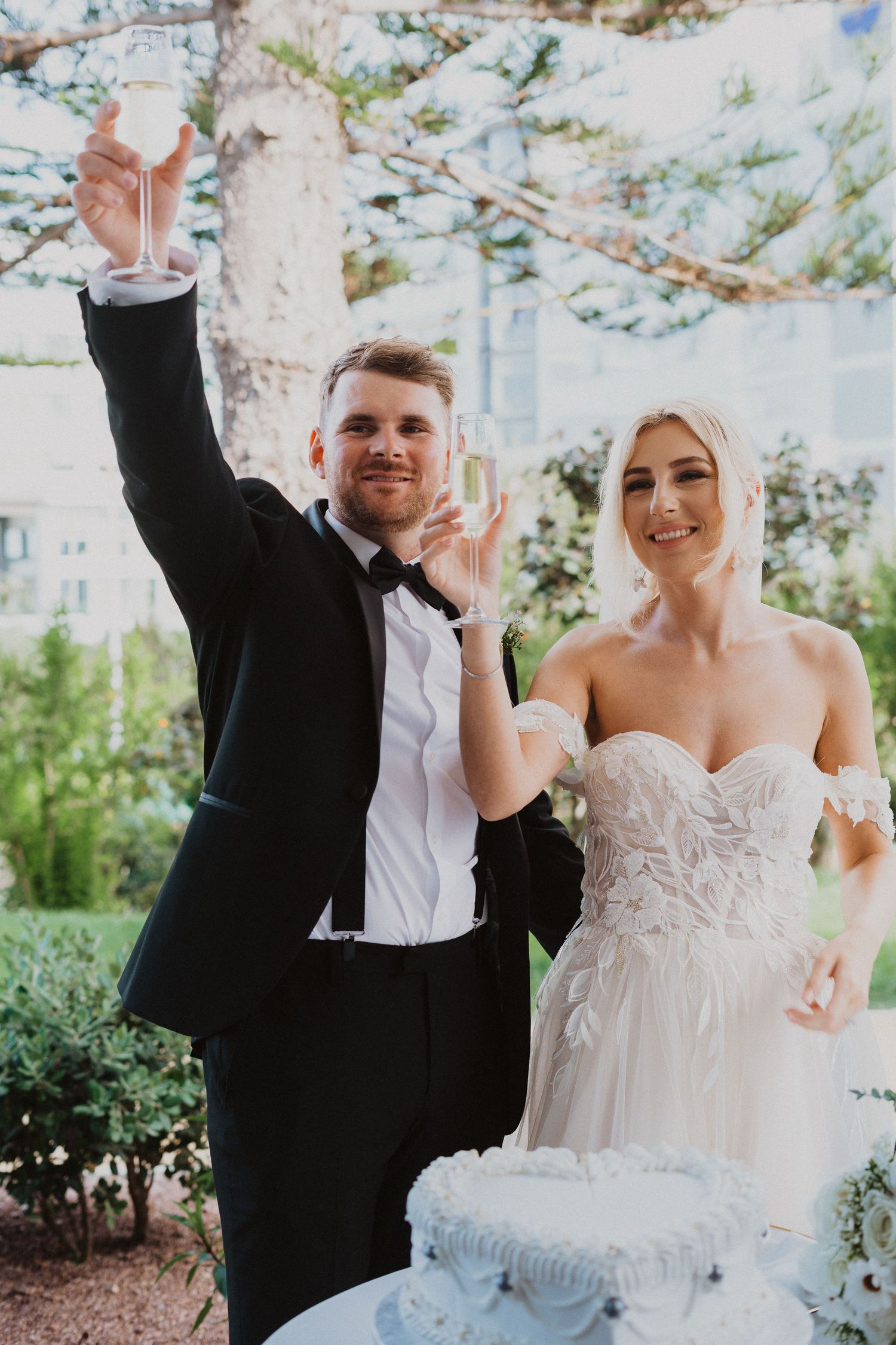 Joyful newlyweds raising champagne glasses in celebration during their outdoor wedding reception.