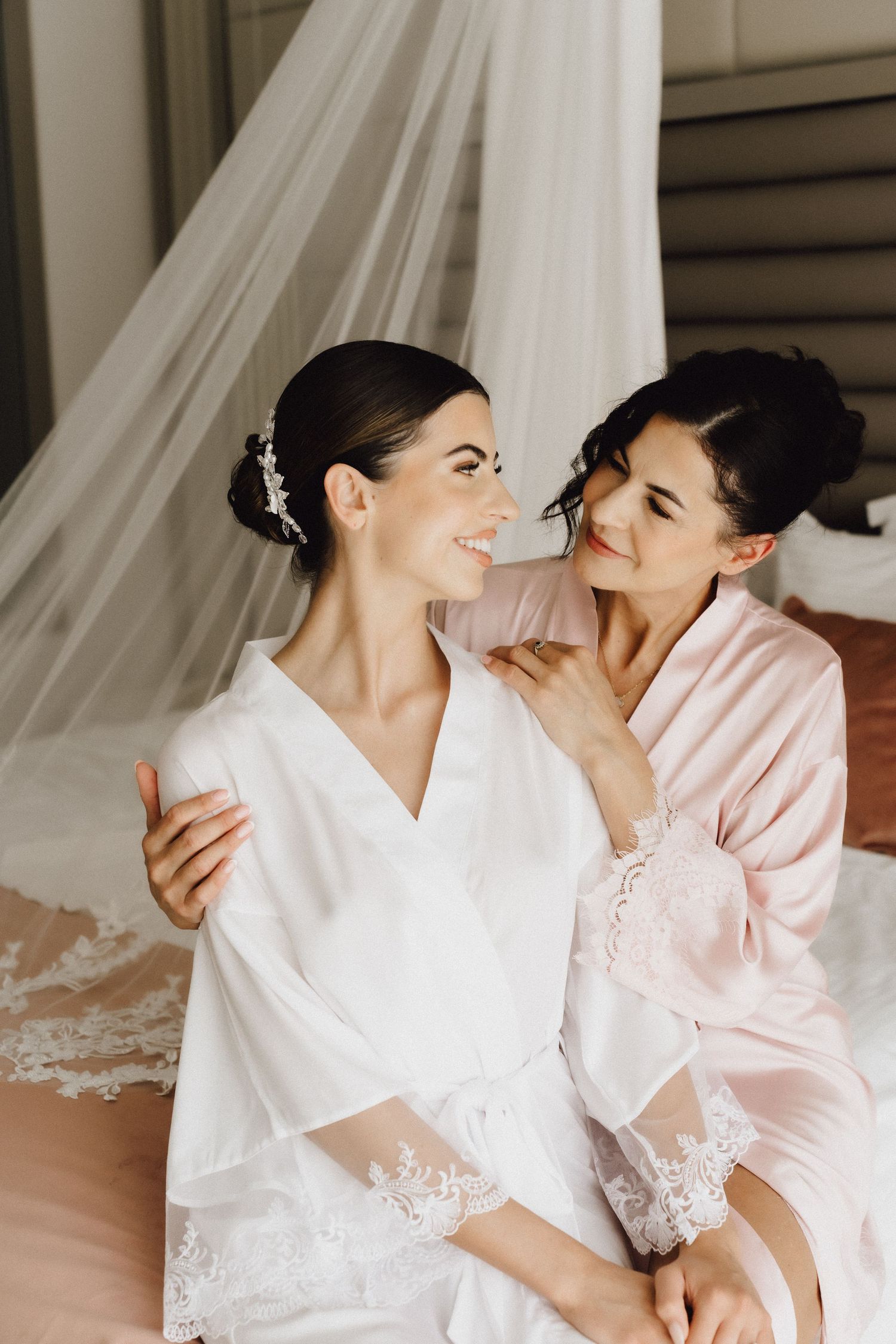 Two people in white and pink robes share a tender moment while sitting on a bed.