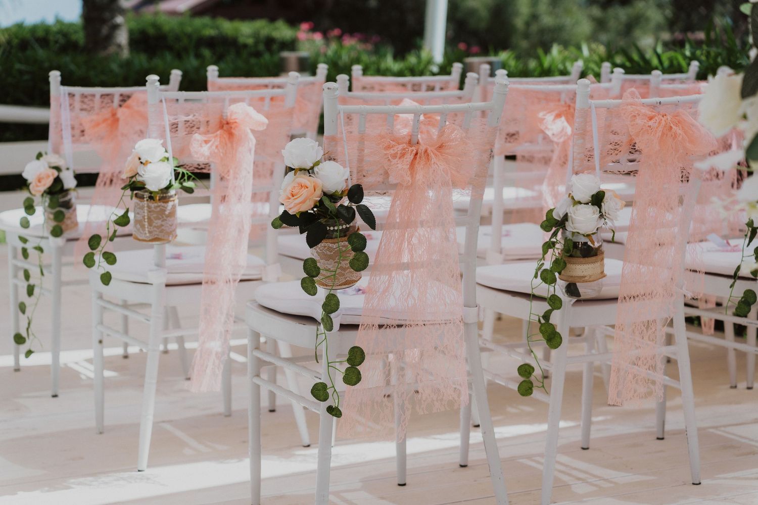 Elegant white wedding chairs decorated with blush pink organza sashes and green floral accents at an outdoor ceremony.