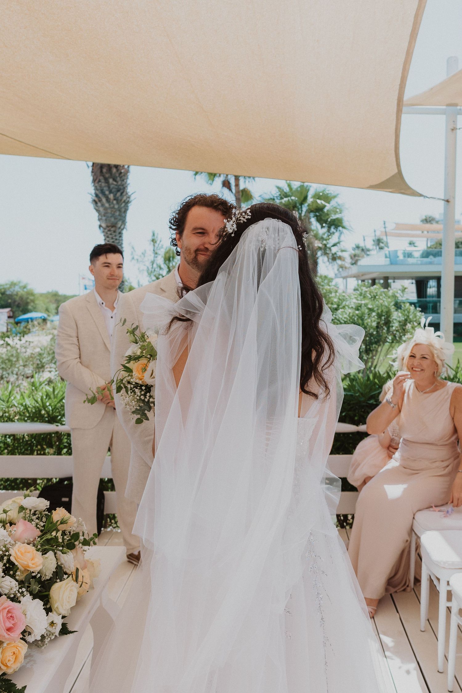 Elegant outdoor wedding ceremony under a beige canopy with floral decorations and seated guests.