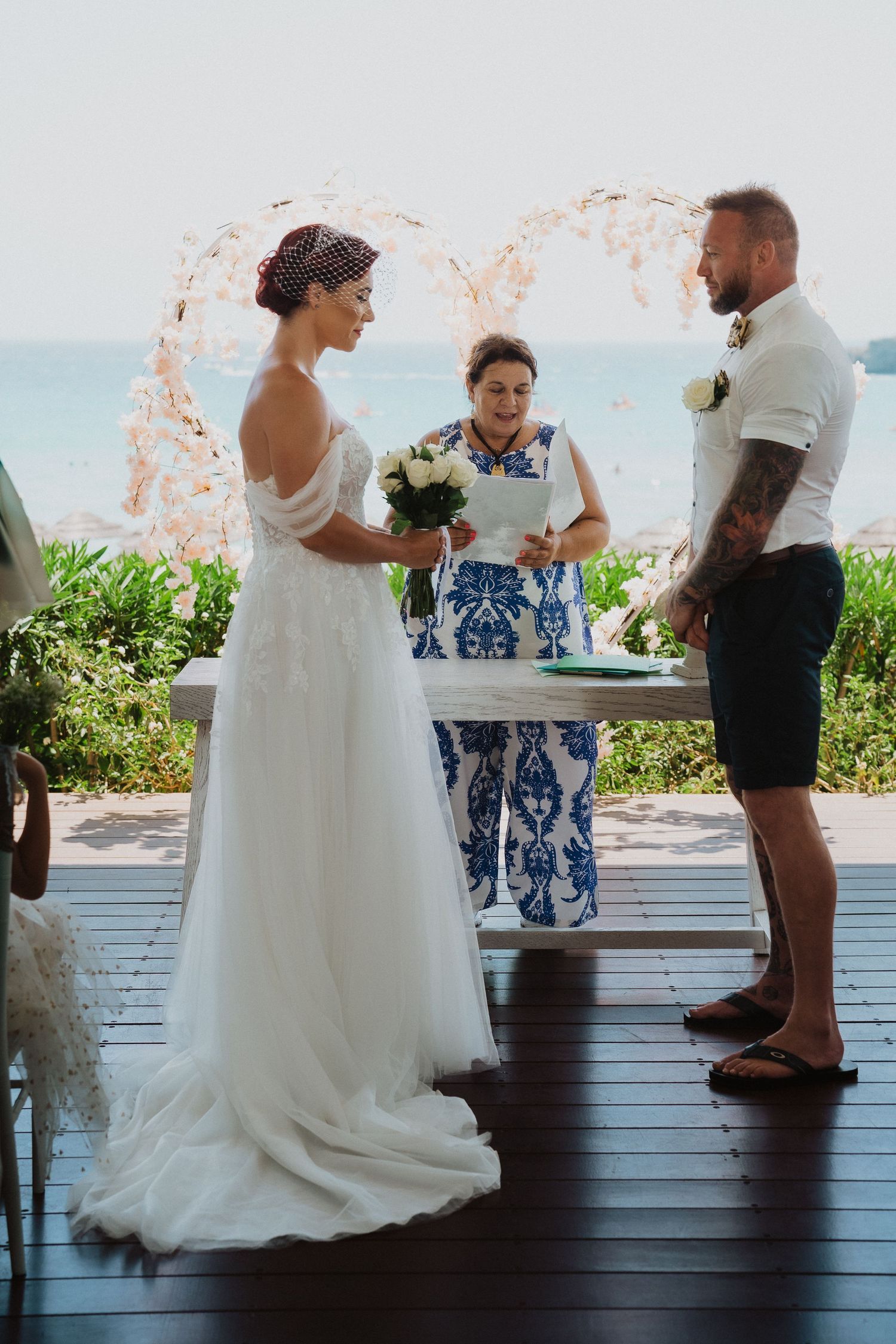 A beach wedding ceremony takes place under a floral arch with the ocean visible in the background.