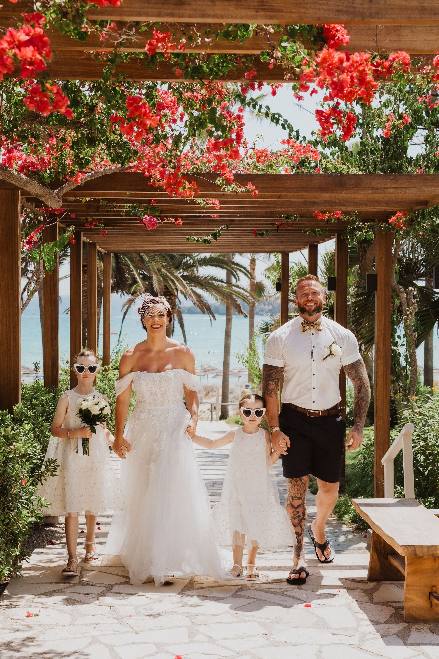 Wedding party walks beneath a flower-covered pergola with ocean views.