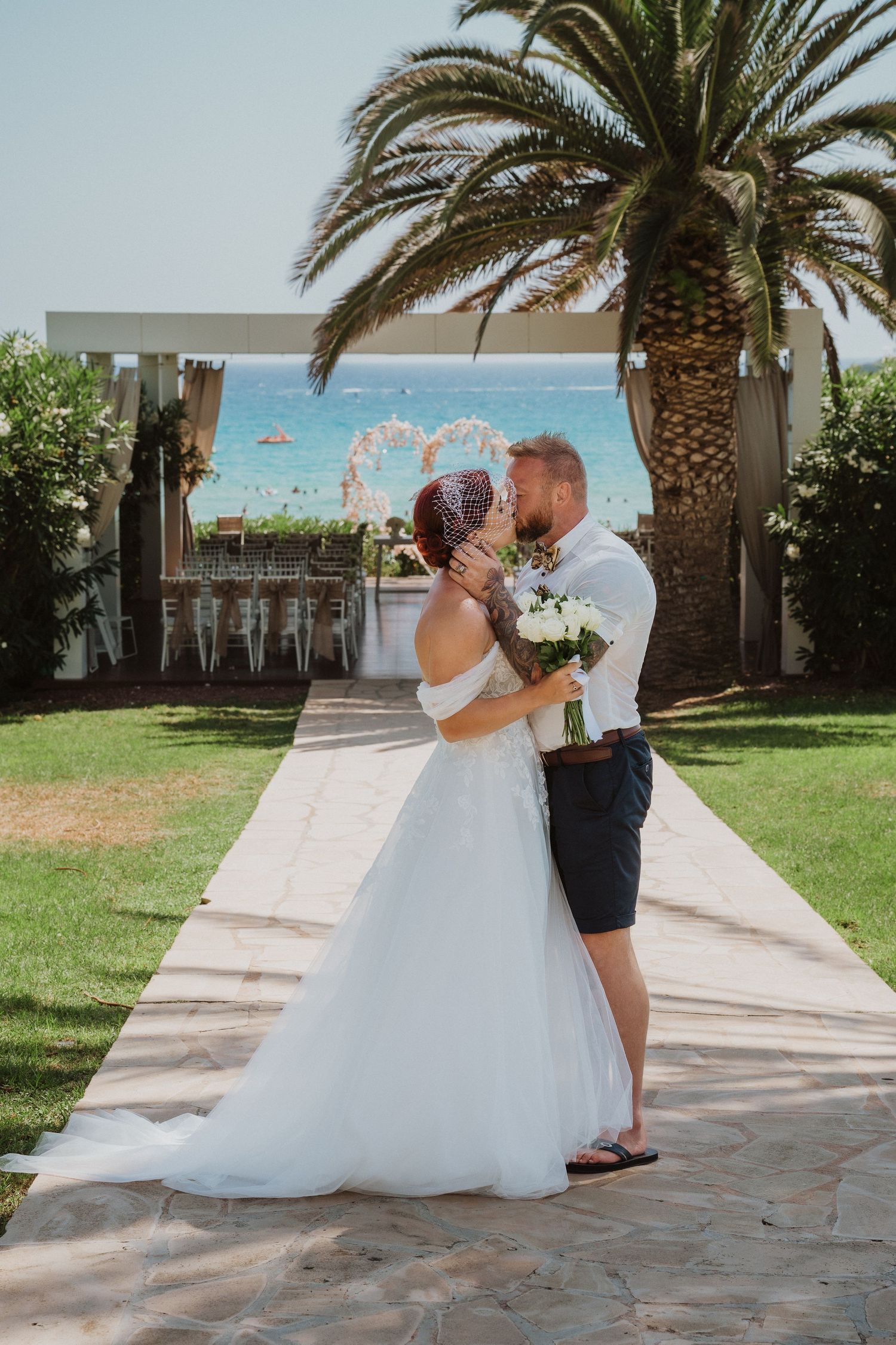 Romantic moment between newlyweds on stone path with palm trees and heart-shaped arch by the ocean.