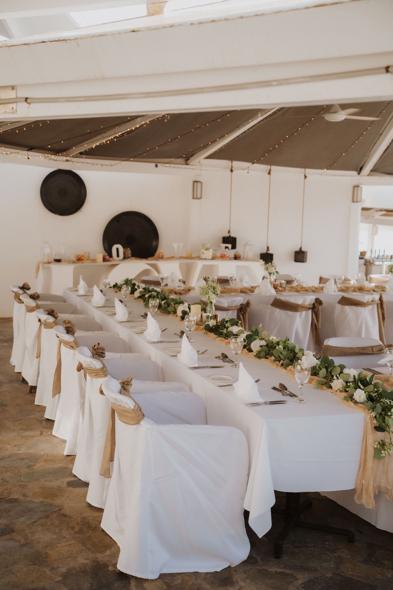 Long banquet table with white linens and greenery garland in bright reception venue.