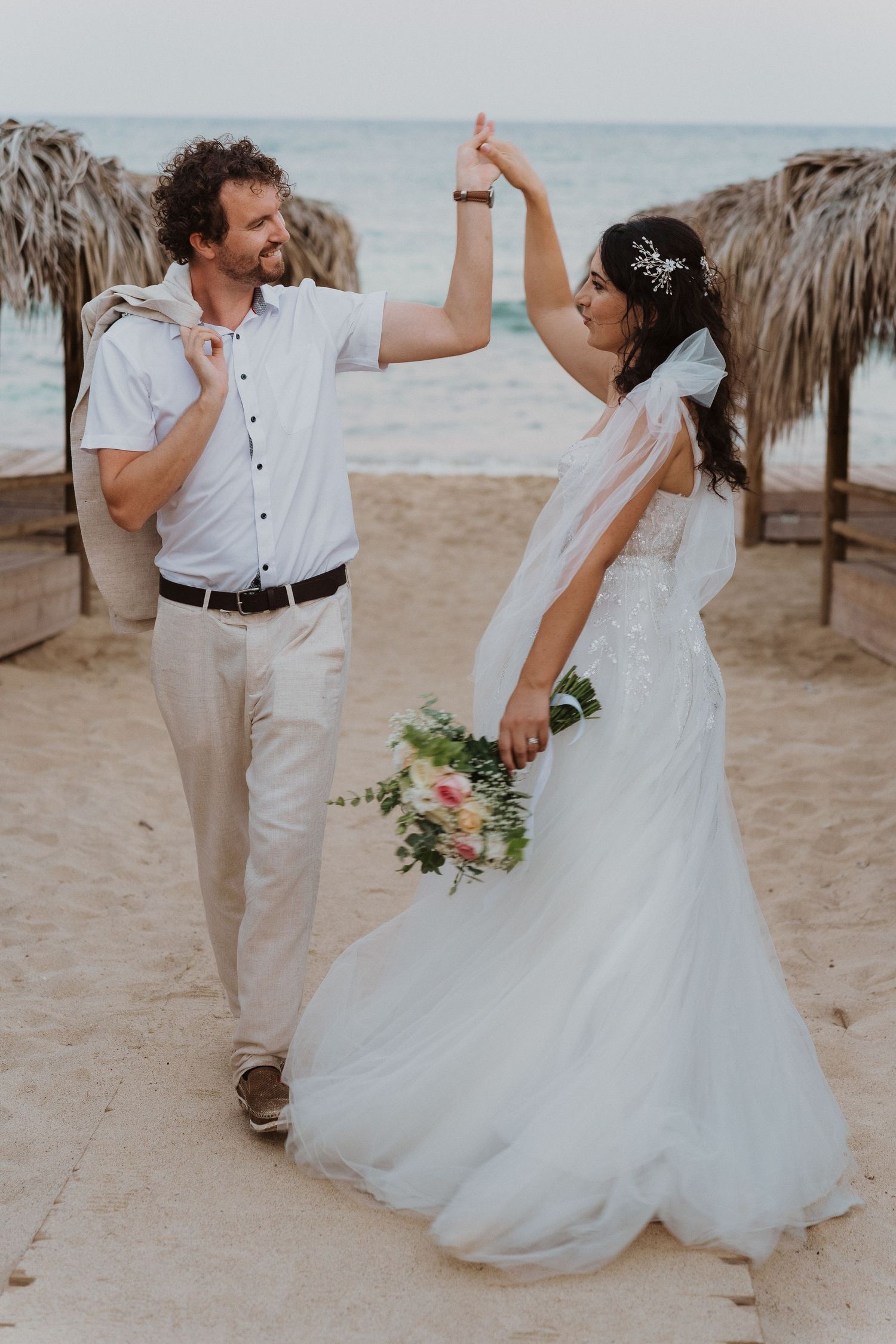 A celebratory wedding moment as a couple dances on the beach under palm frond umbrellas.