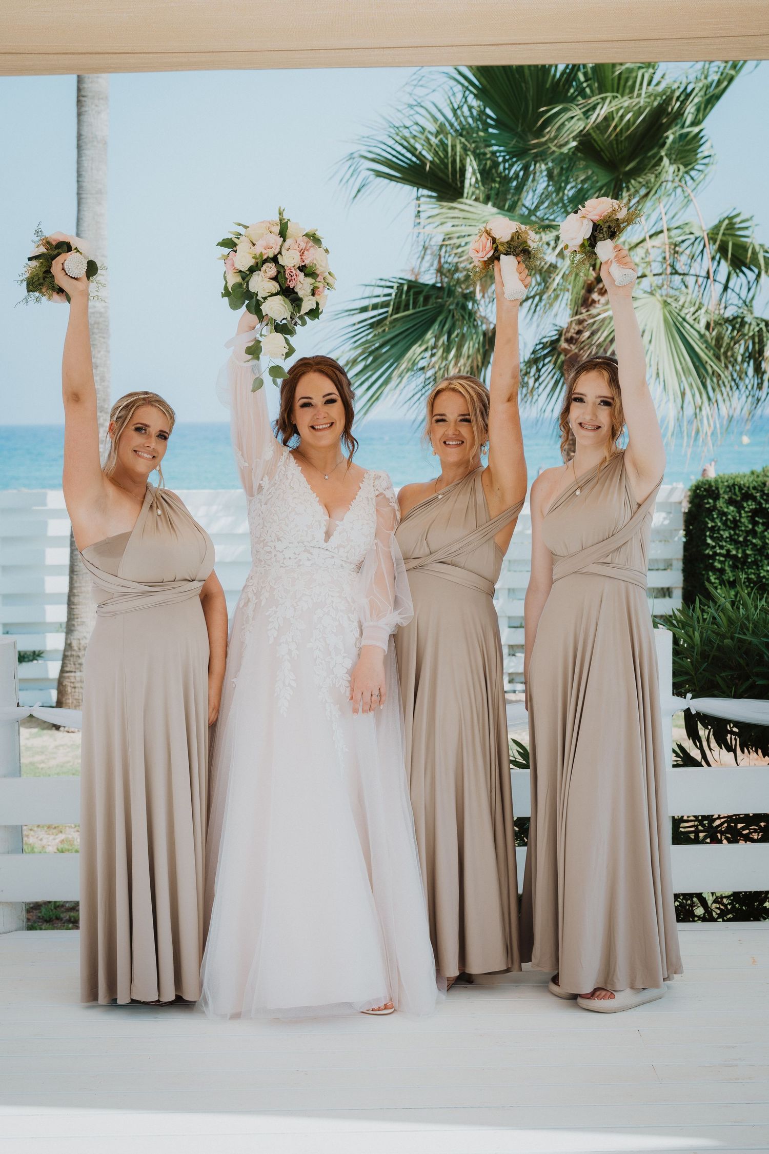Wedding party in beige bridesmaid dresses and white bridal gown pose with raised bouquets by the beach.
