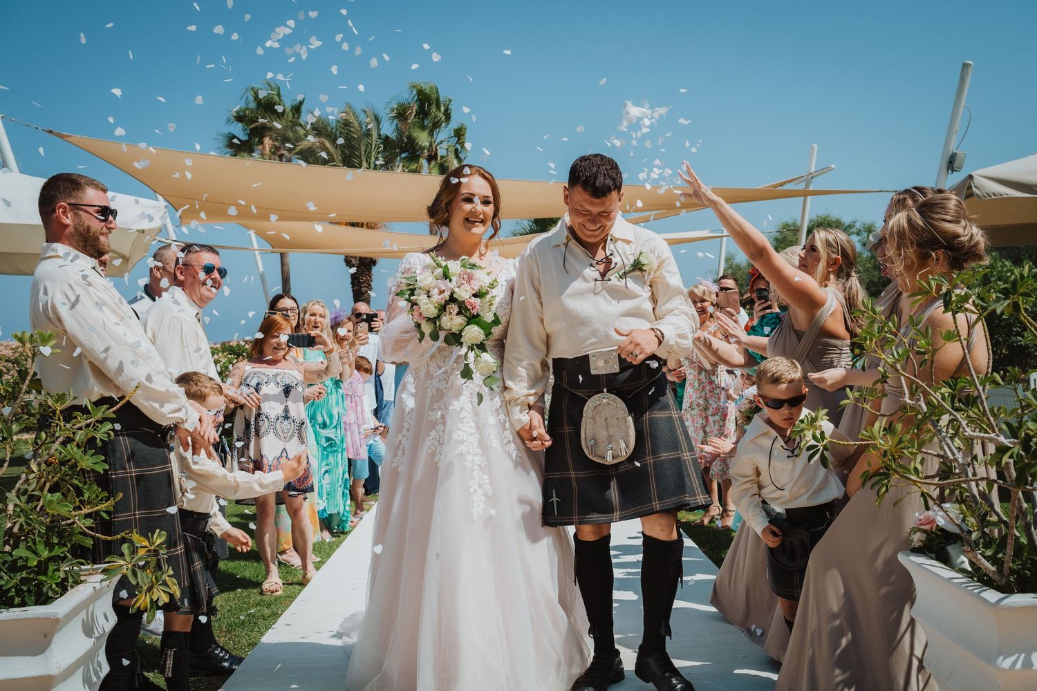 Wedding guests toss confetti at couple during outdoor ceremony with palm trees visible.