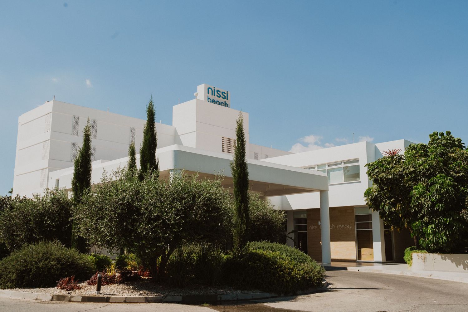 Modern white medical building exterior with tall cypress trees and manicured landscaping under blue sky.