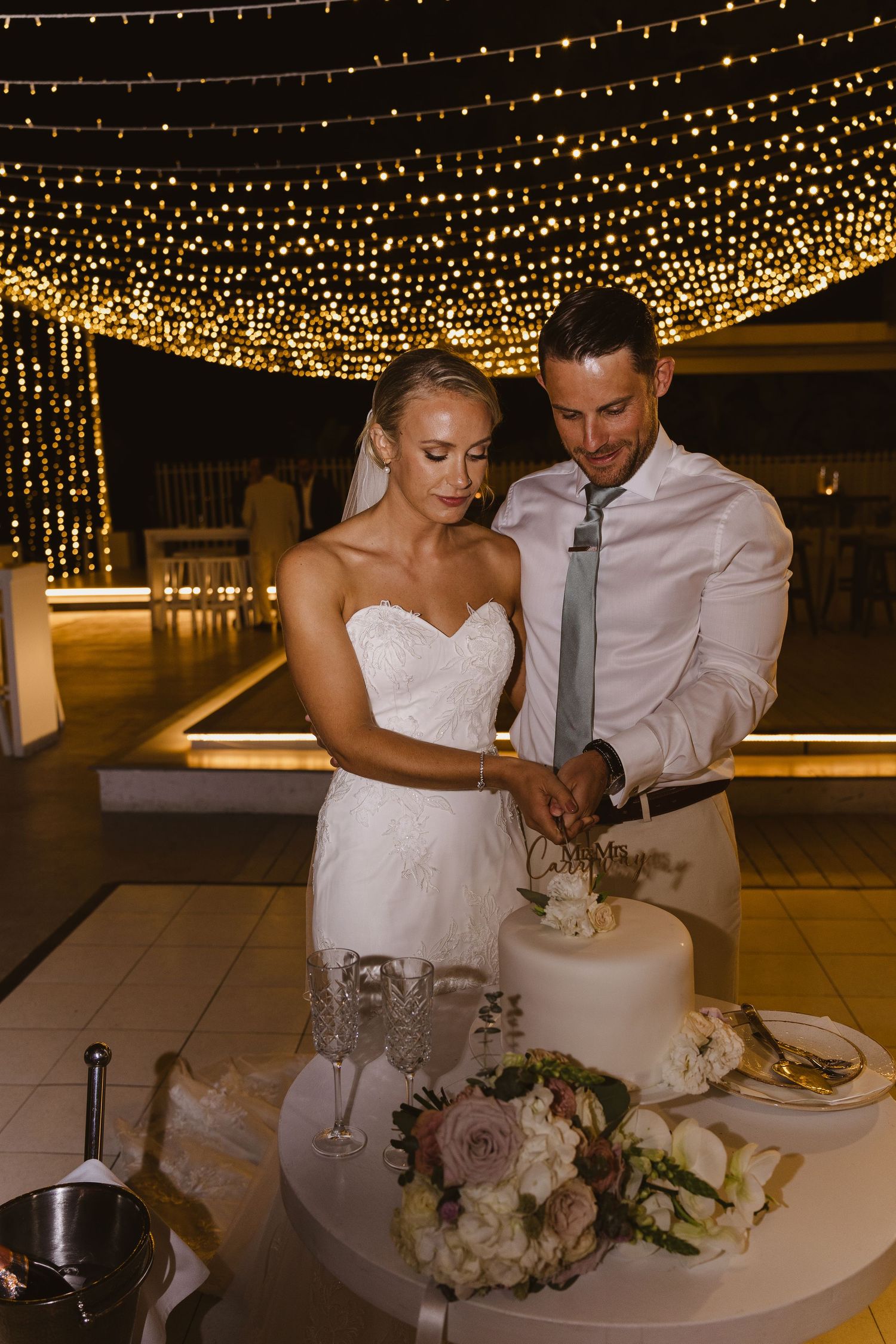 A couple cuts into their wedding cake beneath a canopy of twinkling lights at their reception.