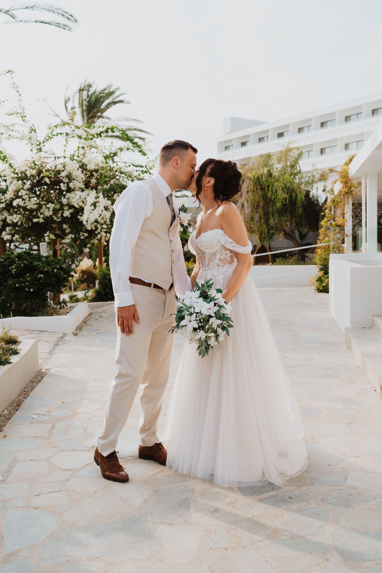 Beautiful destination wedding couple share a romantic moment on a sunny terrace with palm trees and white architecture.