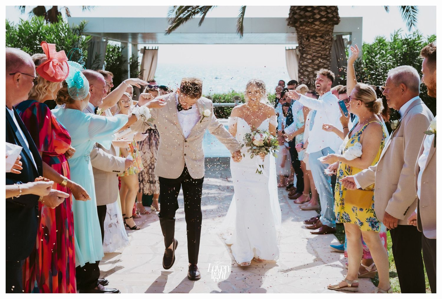Wedding celebration with guests throwing confetti along a palm tree lined walkway.