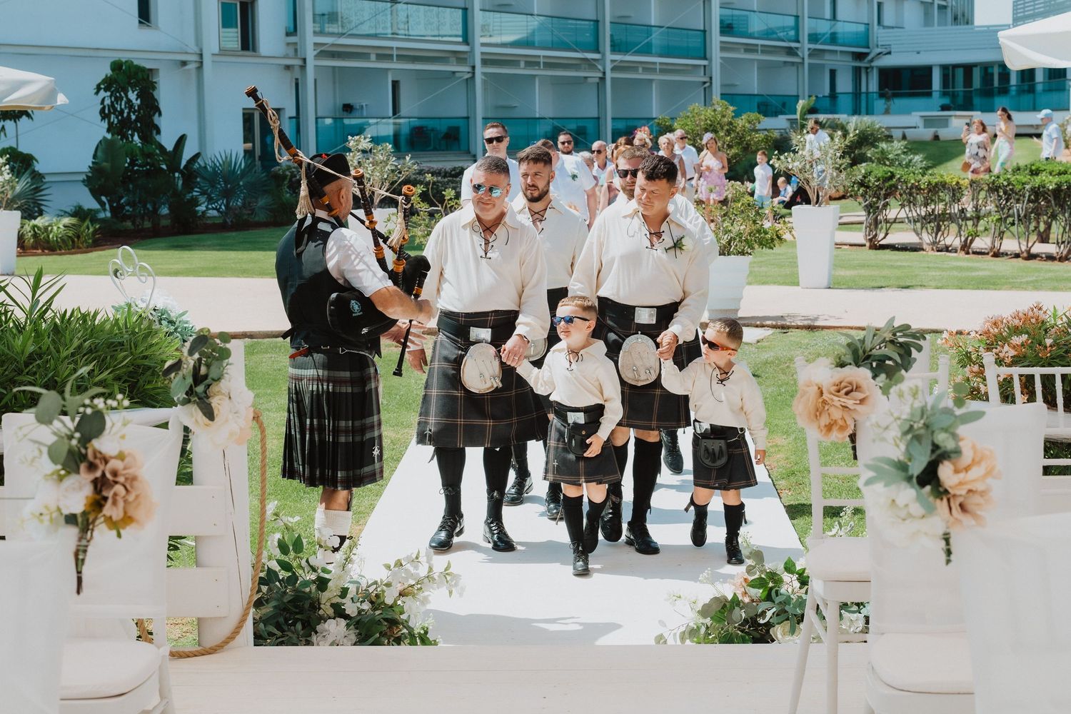 Scottish wedding party in traditional kilts walking down decorated outdoor aisle.