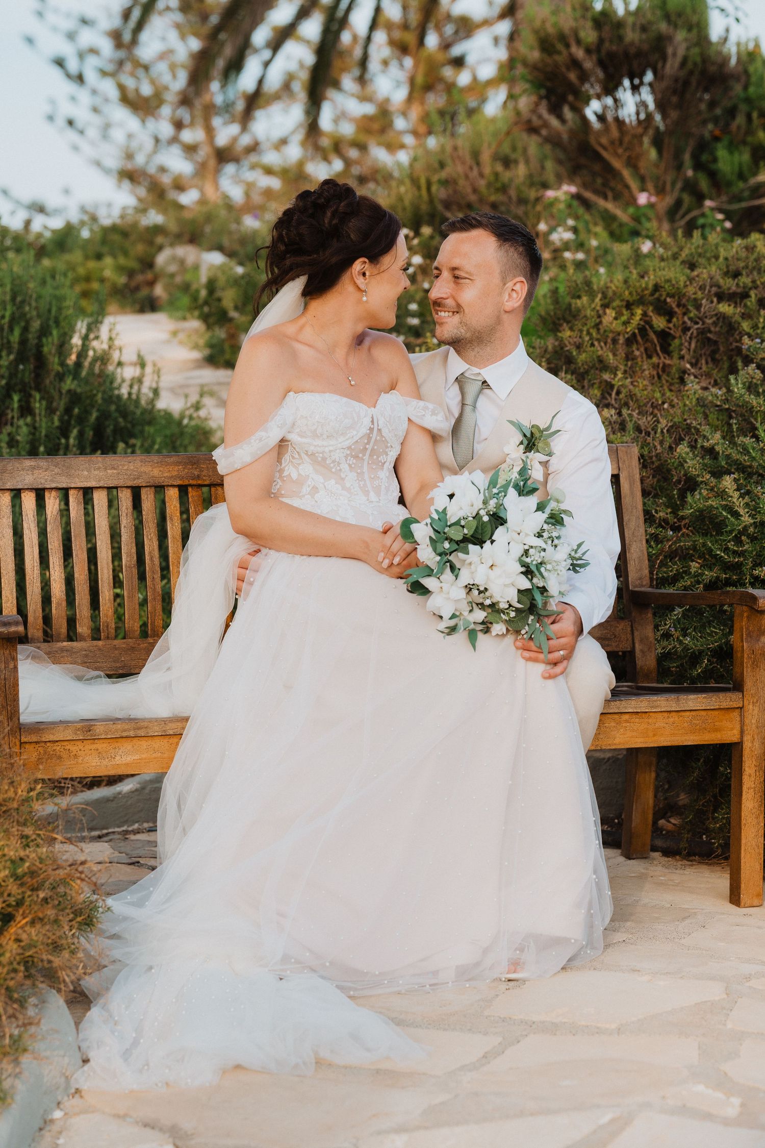 A couple in wedding attire share an intimate moment while sitting on a wooden bench in an outdoor garden setting.