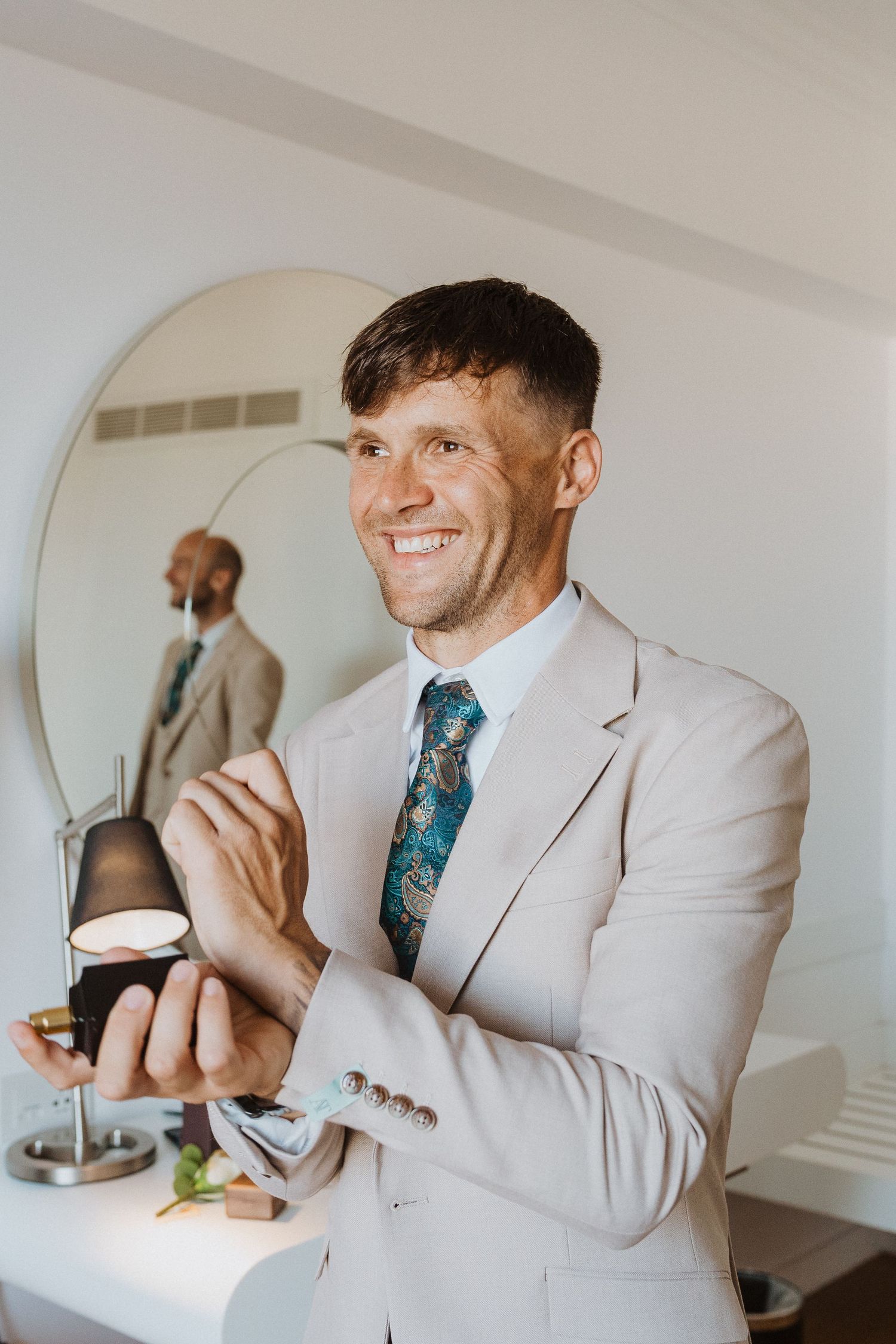 Person in light suit and patterned tie smiling while getting ready in front of circular mirror.