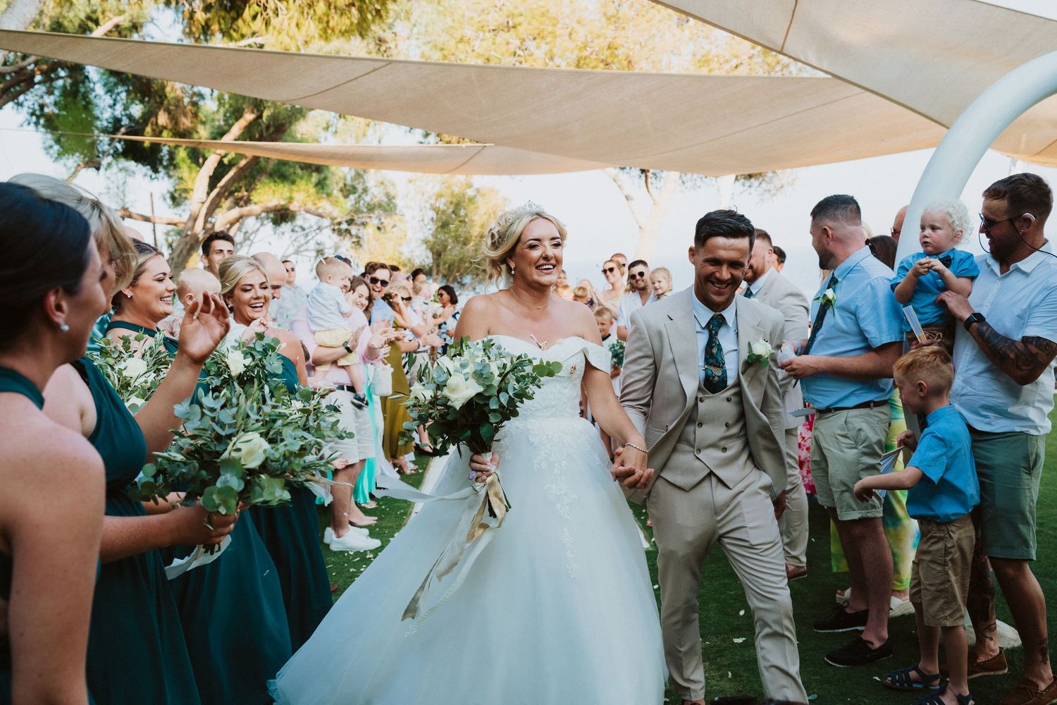 Joyous wedding celebration at an outdoor ceremony with wedding party in teal dresses and light gray suit under white canopy.