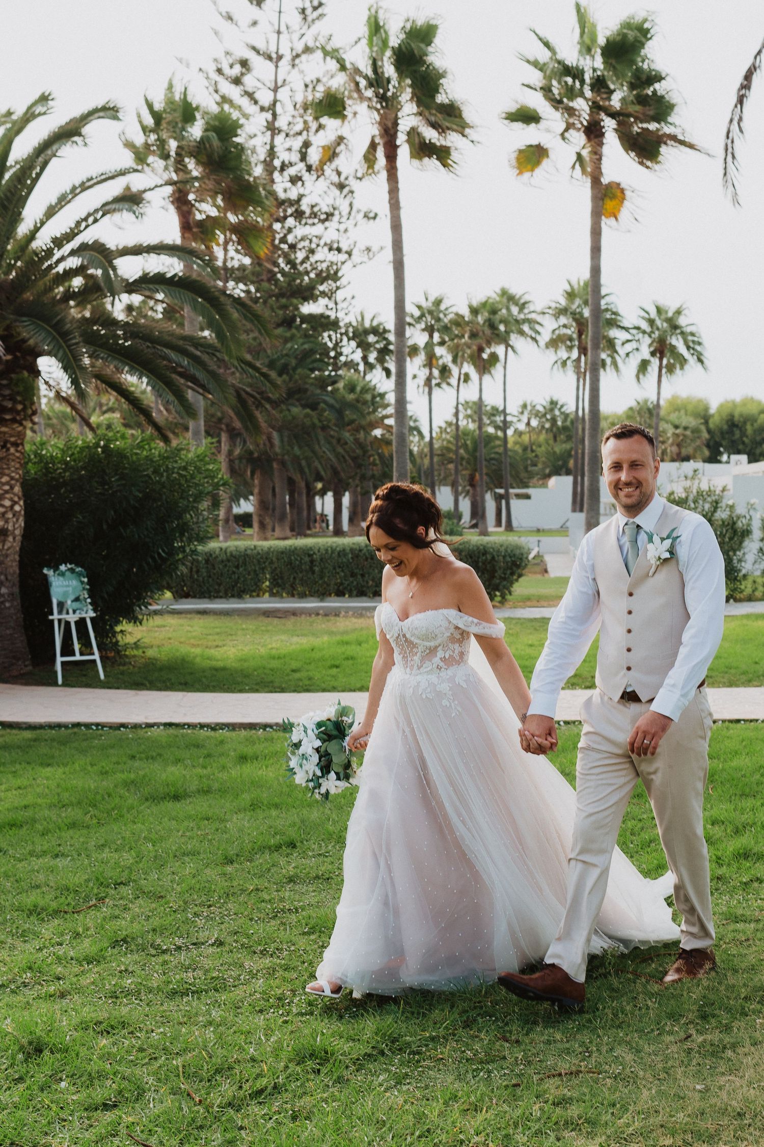 A bride and groom walk hand in hand on a lush lawn surrounded by tall palm trees at an outdoor wedding venue.