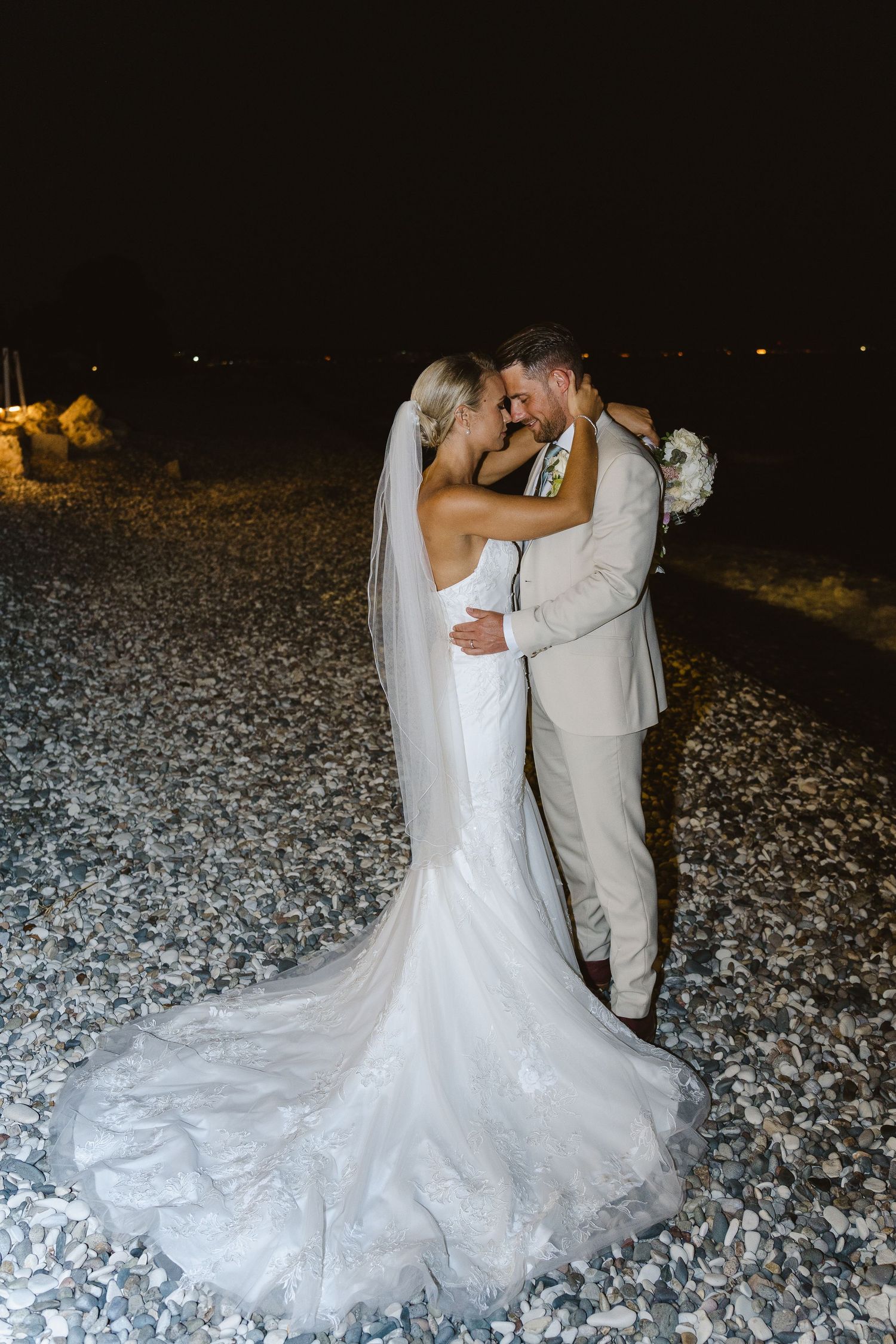 Dramatic nighttime wedding portrait with flowing dress train on rocky beach.