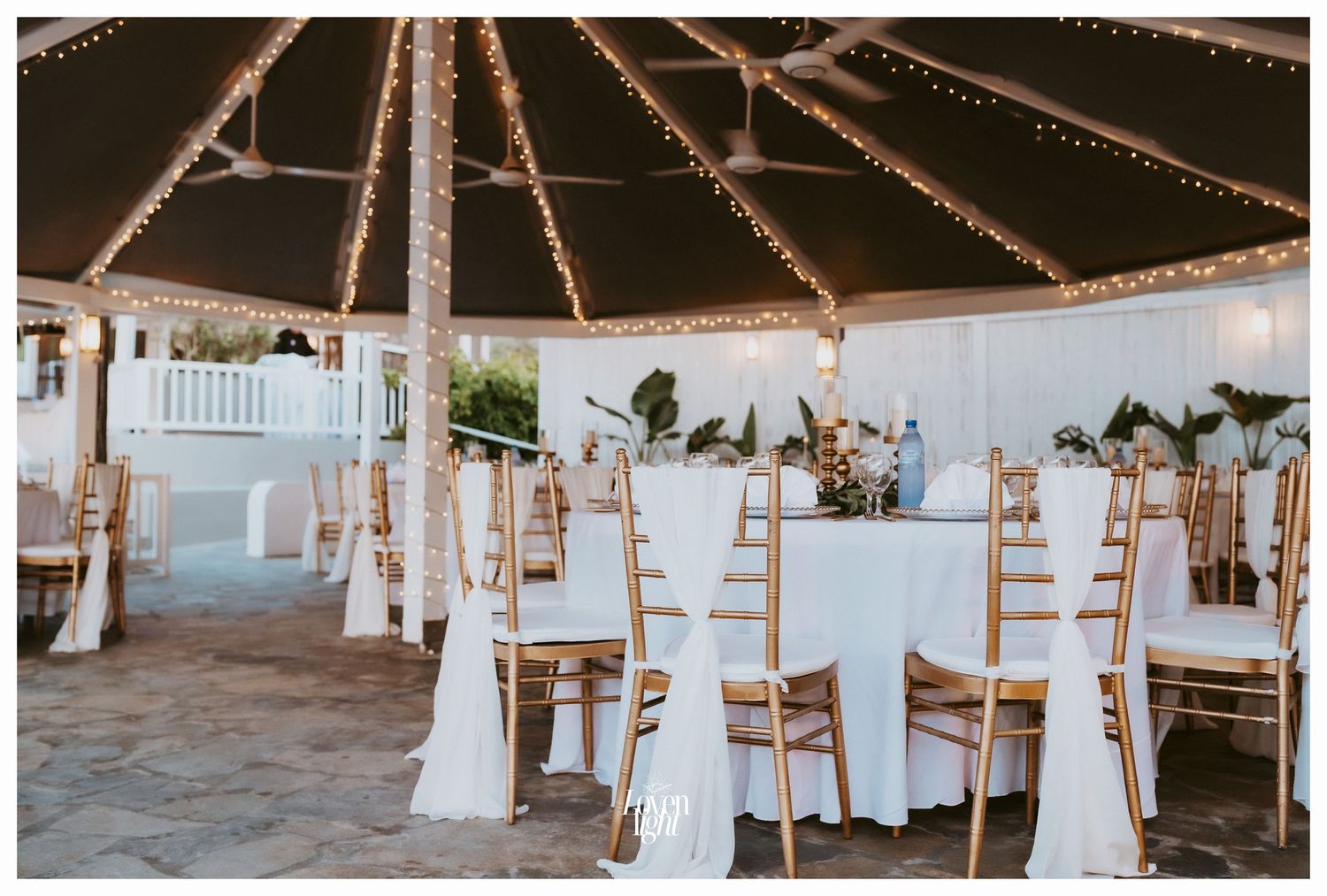 Romantic wedding reception setup with string lights and white-draped chairs under a covered pavilion.